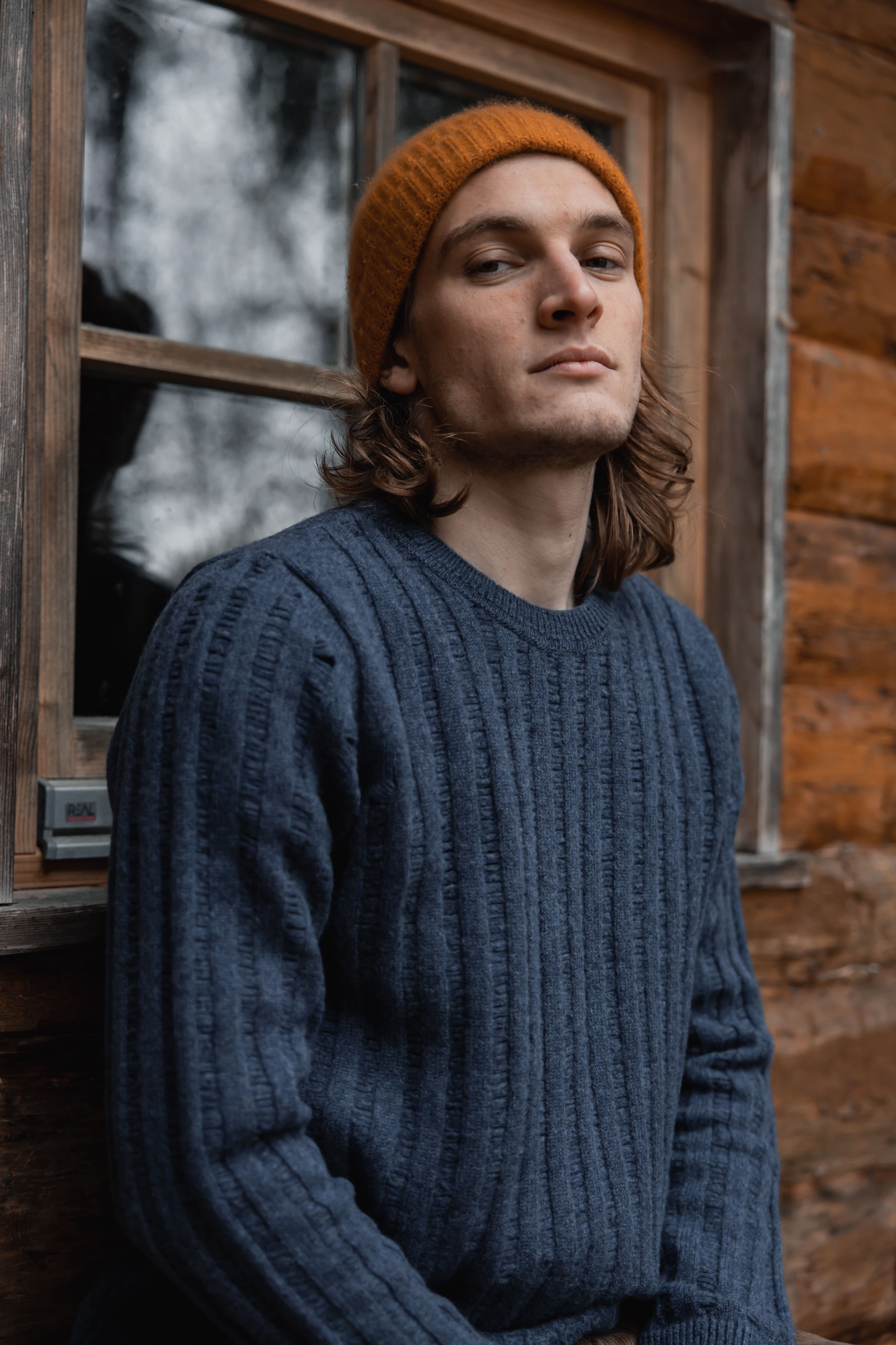 Young man wearing an orange knit beanie and a blue textured sweater leaning against a wooden cabin wall near a window.