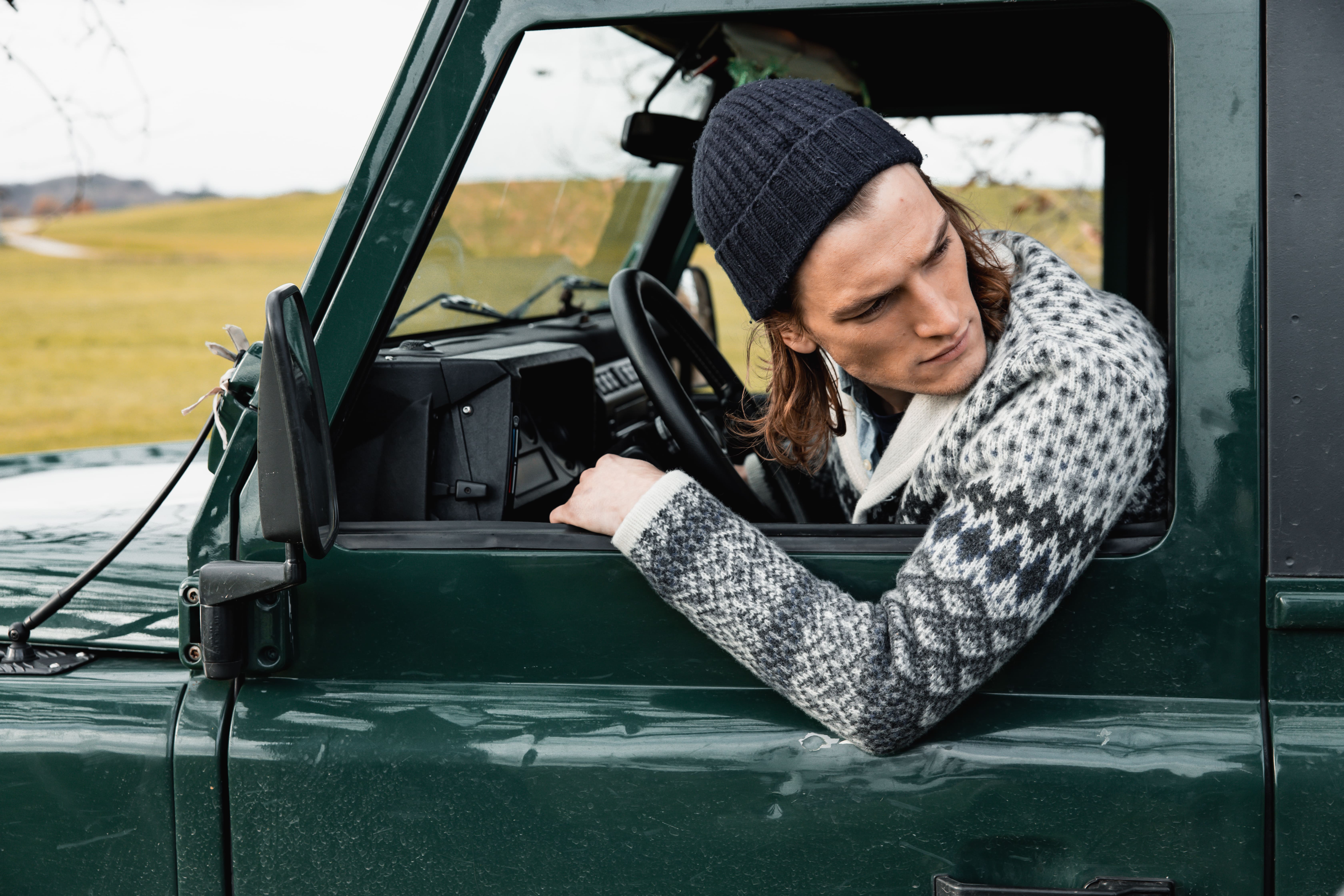 Man wearing a knitted beanie and patterned sweater looking to his left out the window of a green vehicle.