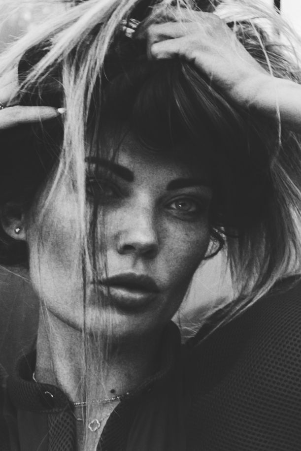 Black and white close-up portrait of a woman with intense eyes, tousled hair, and hands in her hair.