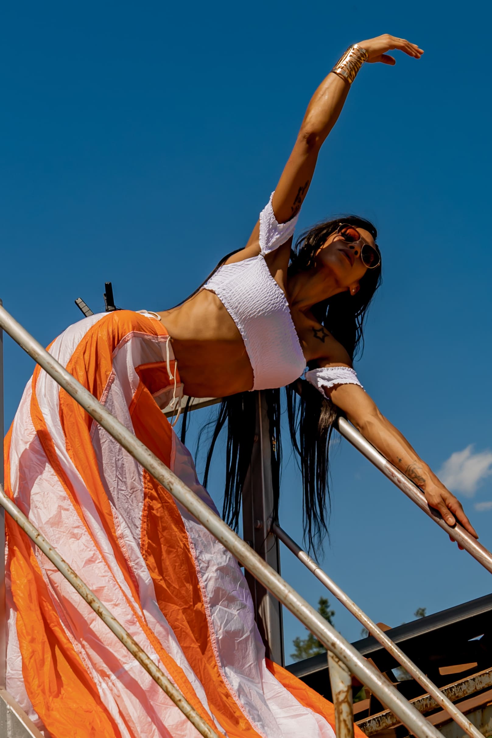 Woman in sunglasses wearing a white cropped top and orange-white billowing skirt, leaning back on metal railings under a clear blue sky.
