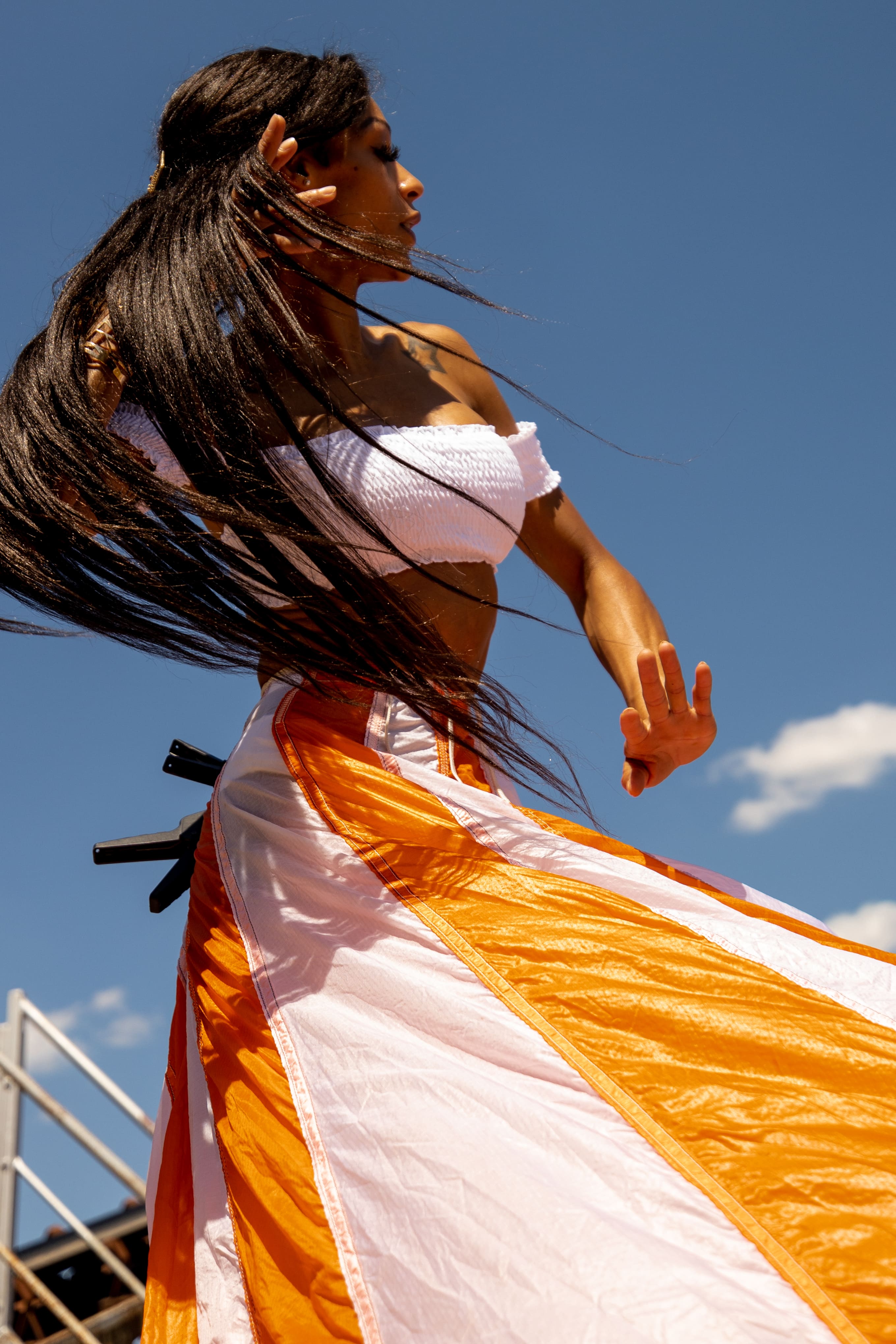 Woman with long dark hair wearing a white crop top and flowing orange and white skirt against a clear blue sky.