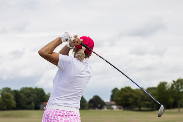 Golfer in pink hat and patterned shorts swinging a driver on a grassy course under a cloudy sky.