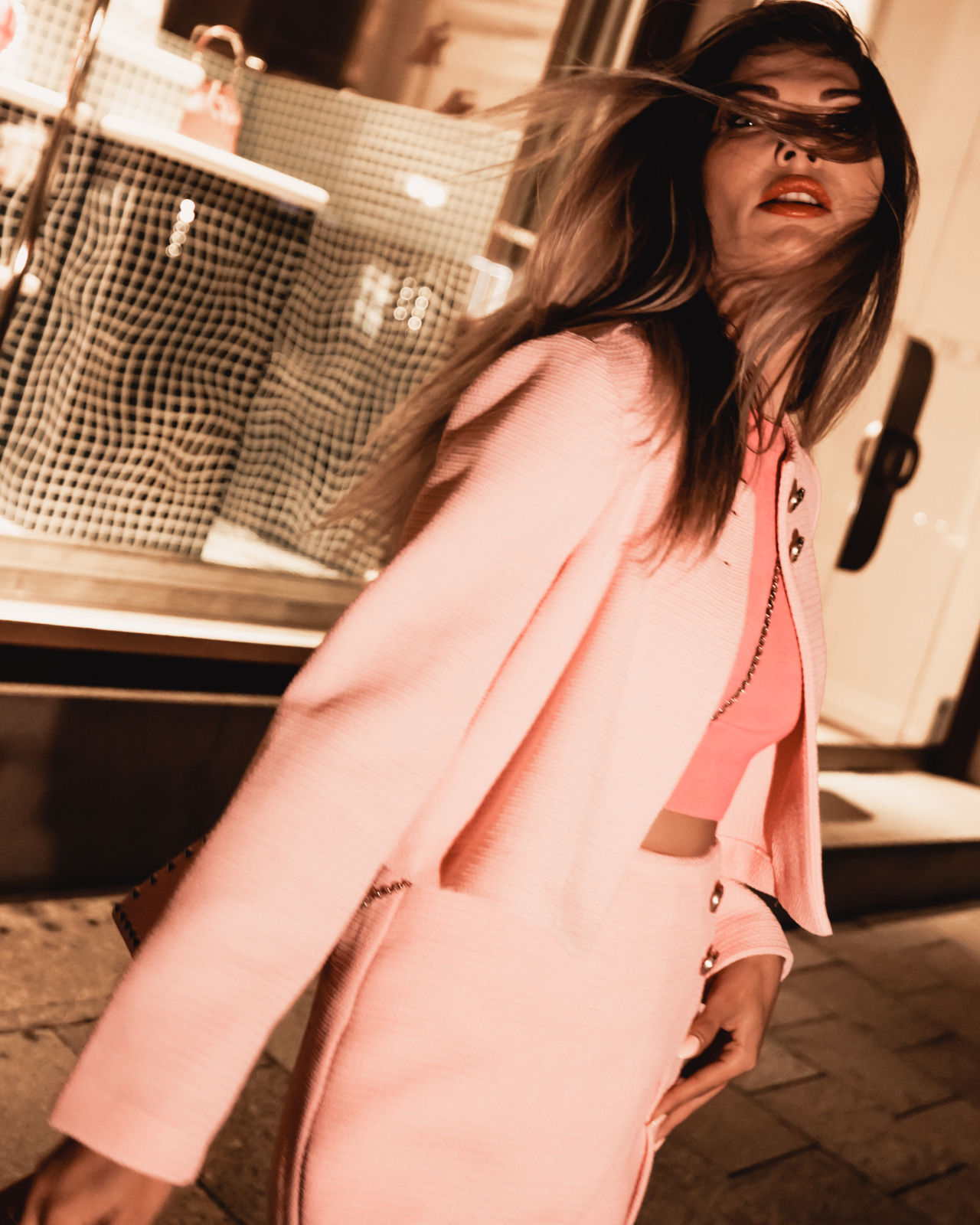 Woman wearing a pink skirt suit with flowing hair and bold red lipstick, walking outdoors at night.