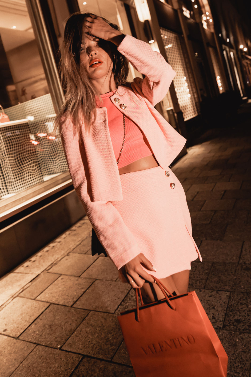Woman in pink outfit holding an orange Valentino shopping bag on a city sidewalk at night.