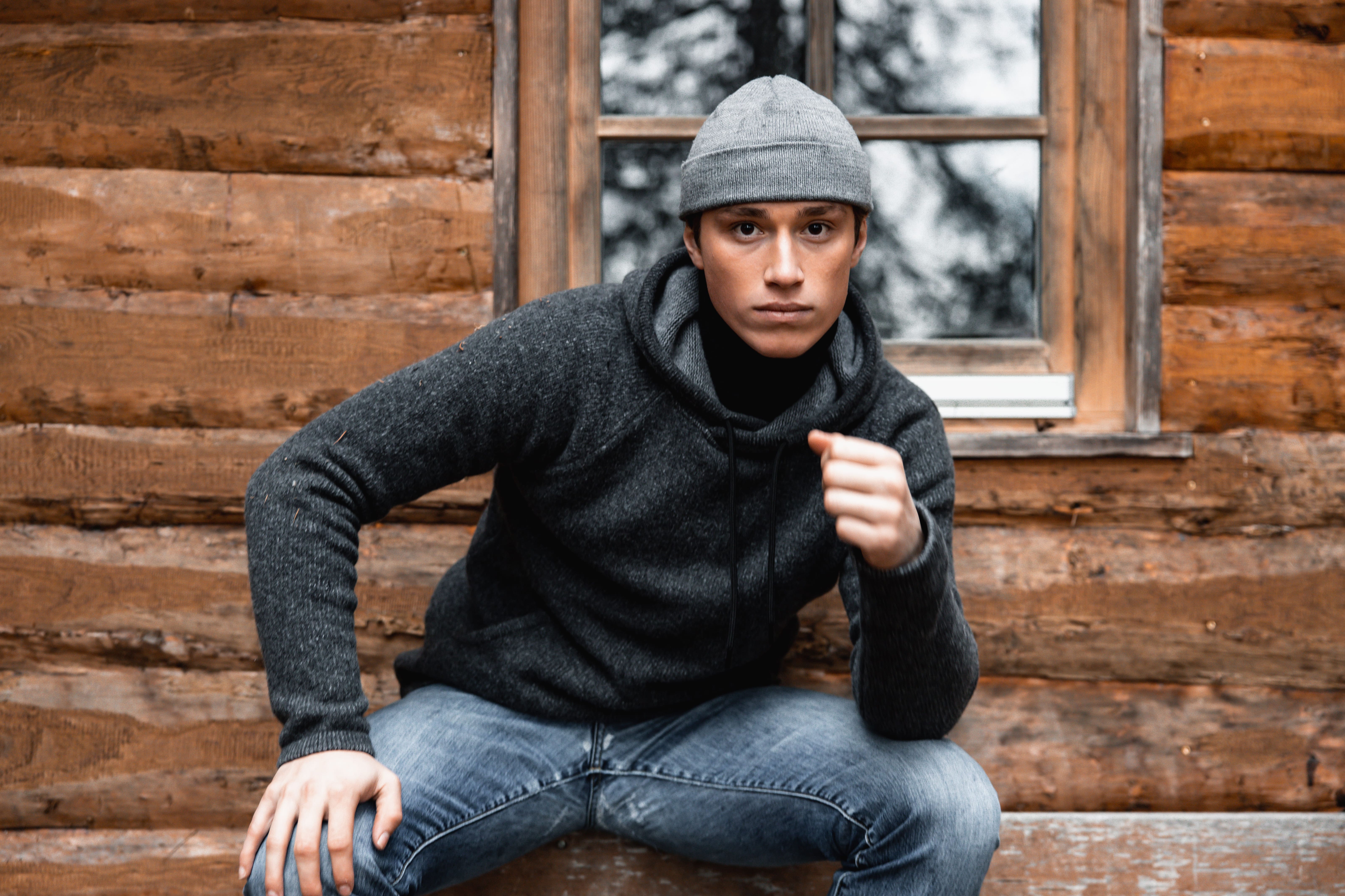 Young man in a gray beanie and dark sweater crouching in front of a wooden cabin with a window.