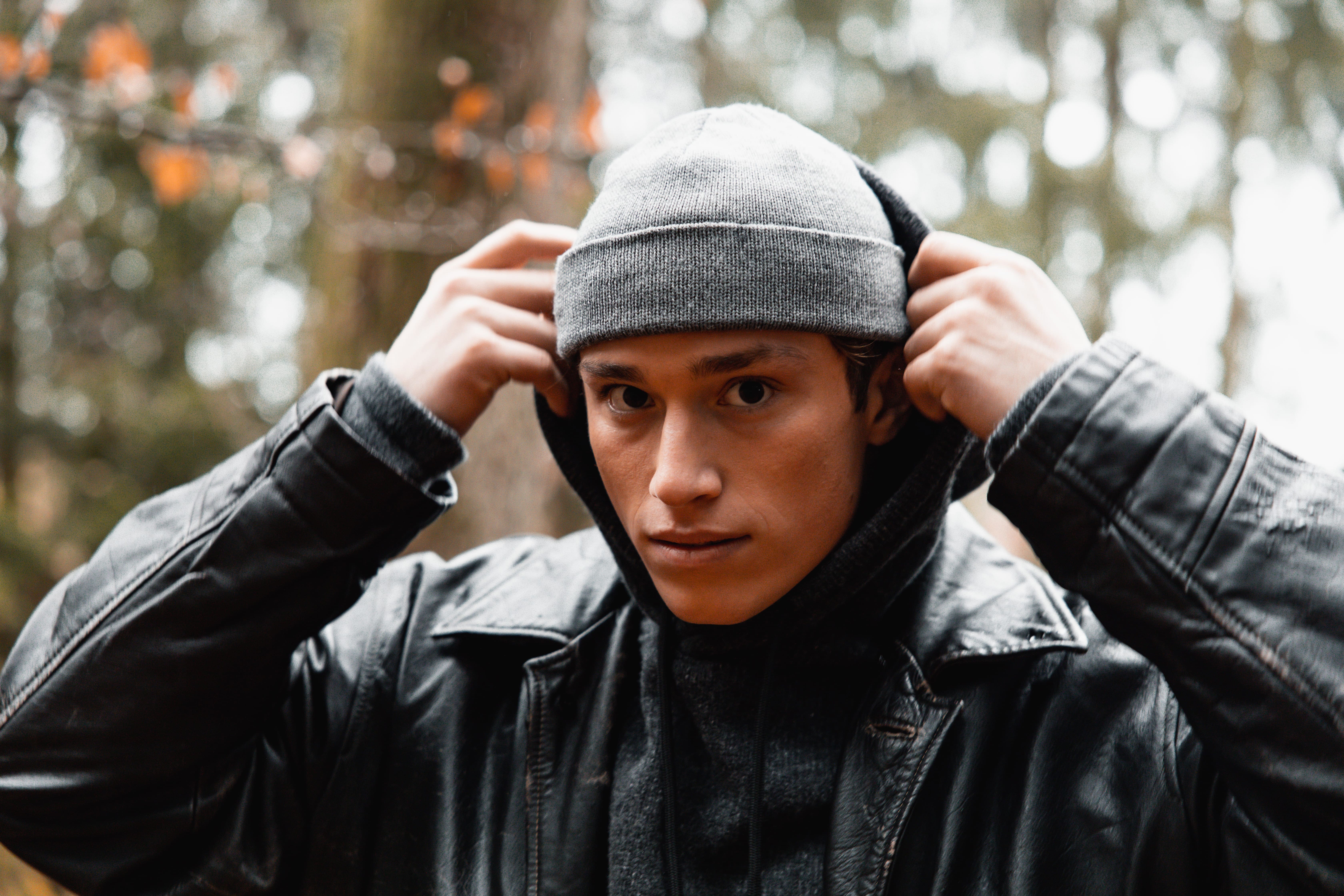 Young man outdoors wearing a gray beanie and black leather jacket, pulling up his hoodie.