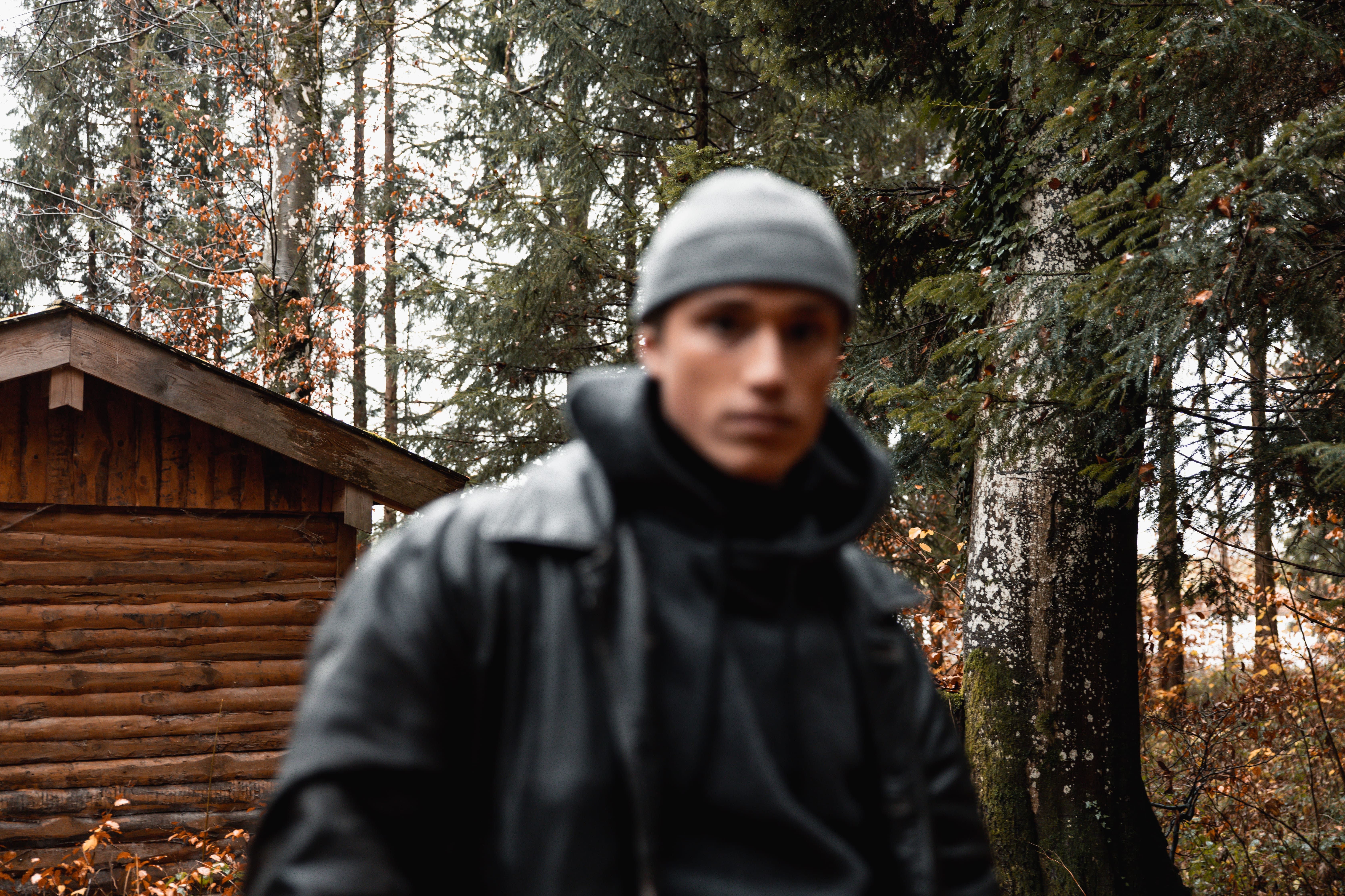 Person wearing a gray beanie and black jacket standing outdoors with a wooden cabin and trees in the background.