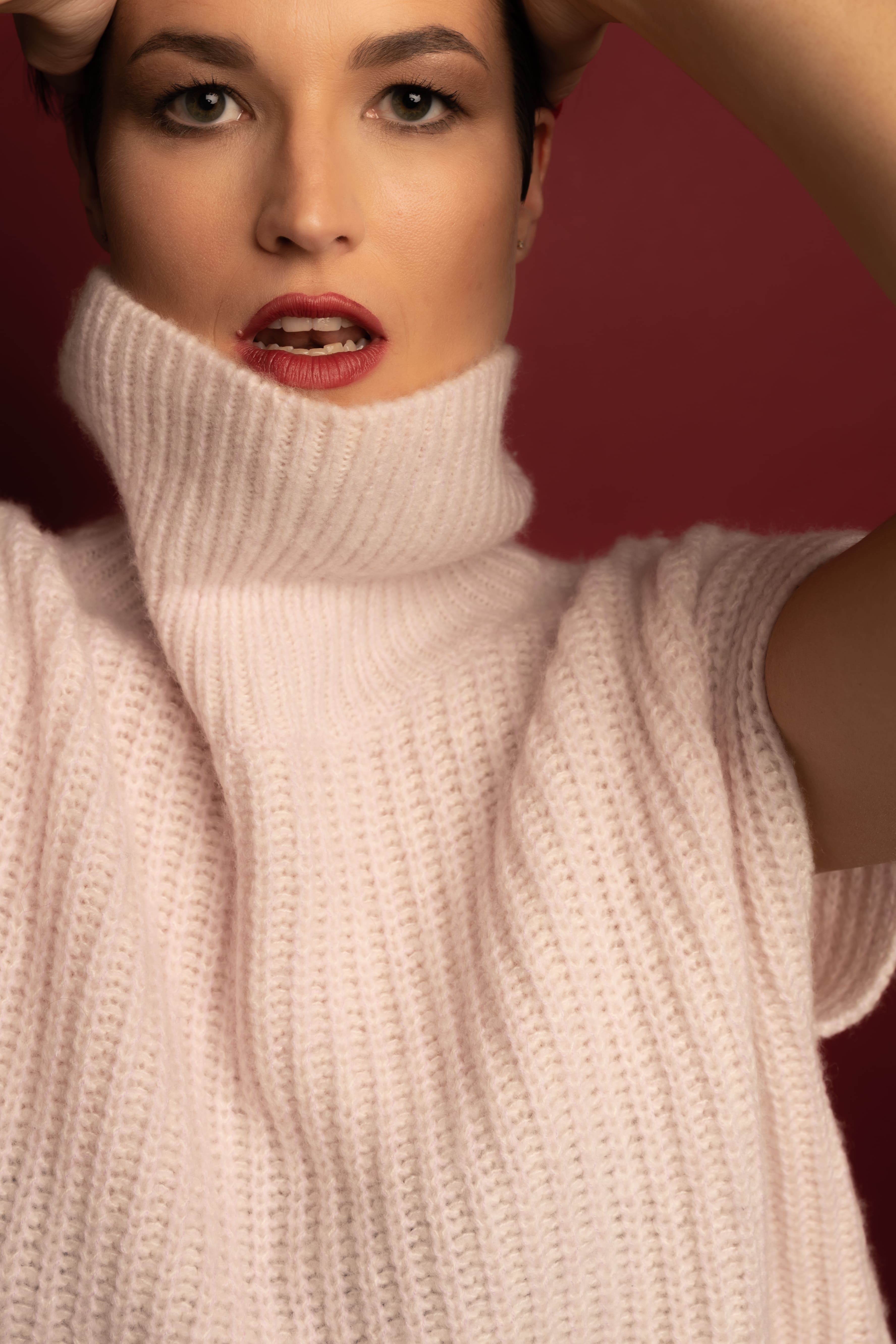 Close-up of a woman with short dark hair wearing a light pink chunky knit turtleneck sweater against a maroon background.