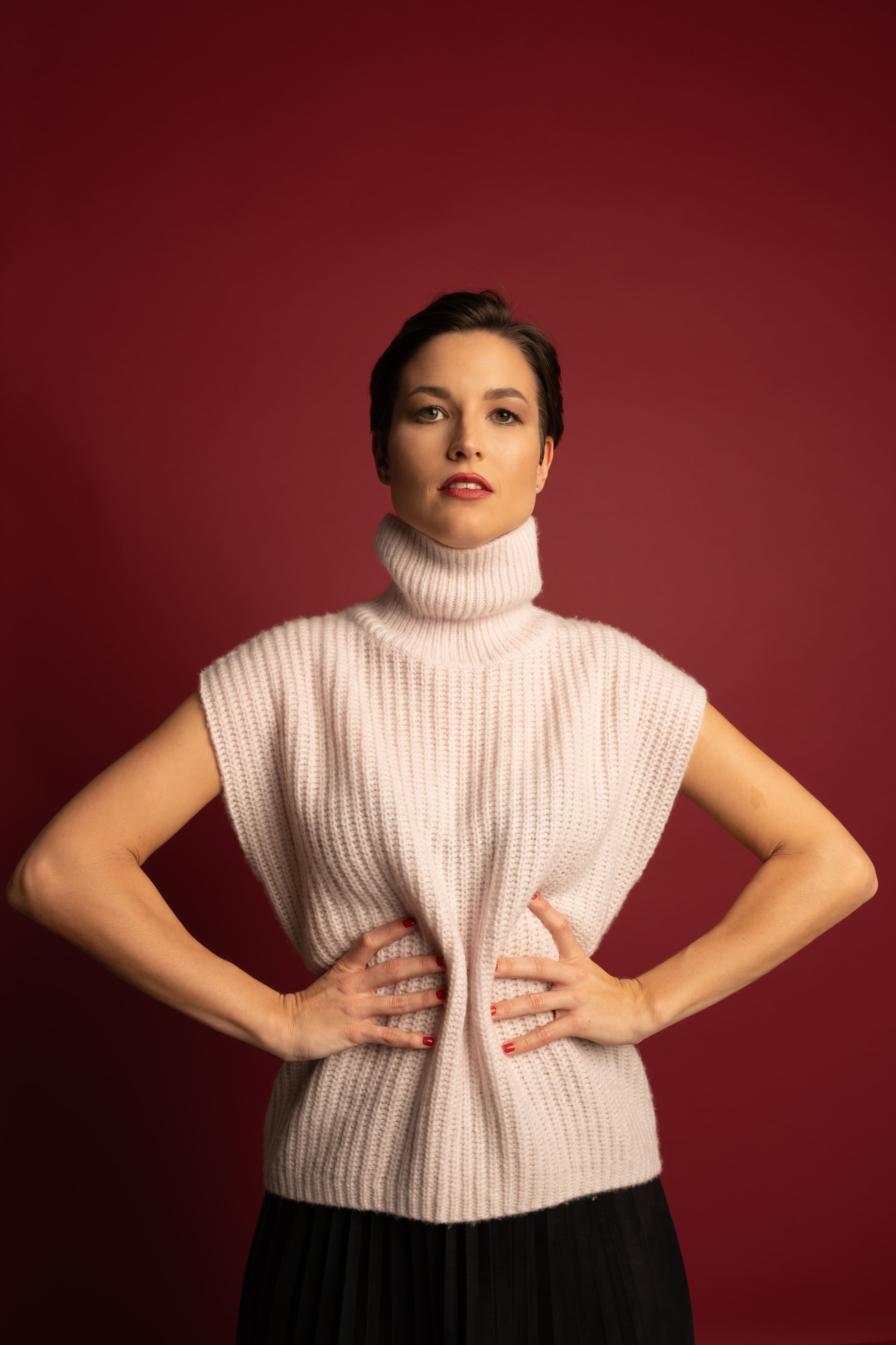 Woman with short dark hair wearing a sleeveless white turtleneck sweater posing with hands on waist against a dark red background.