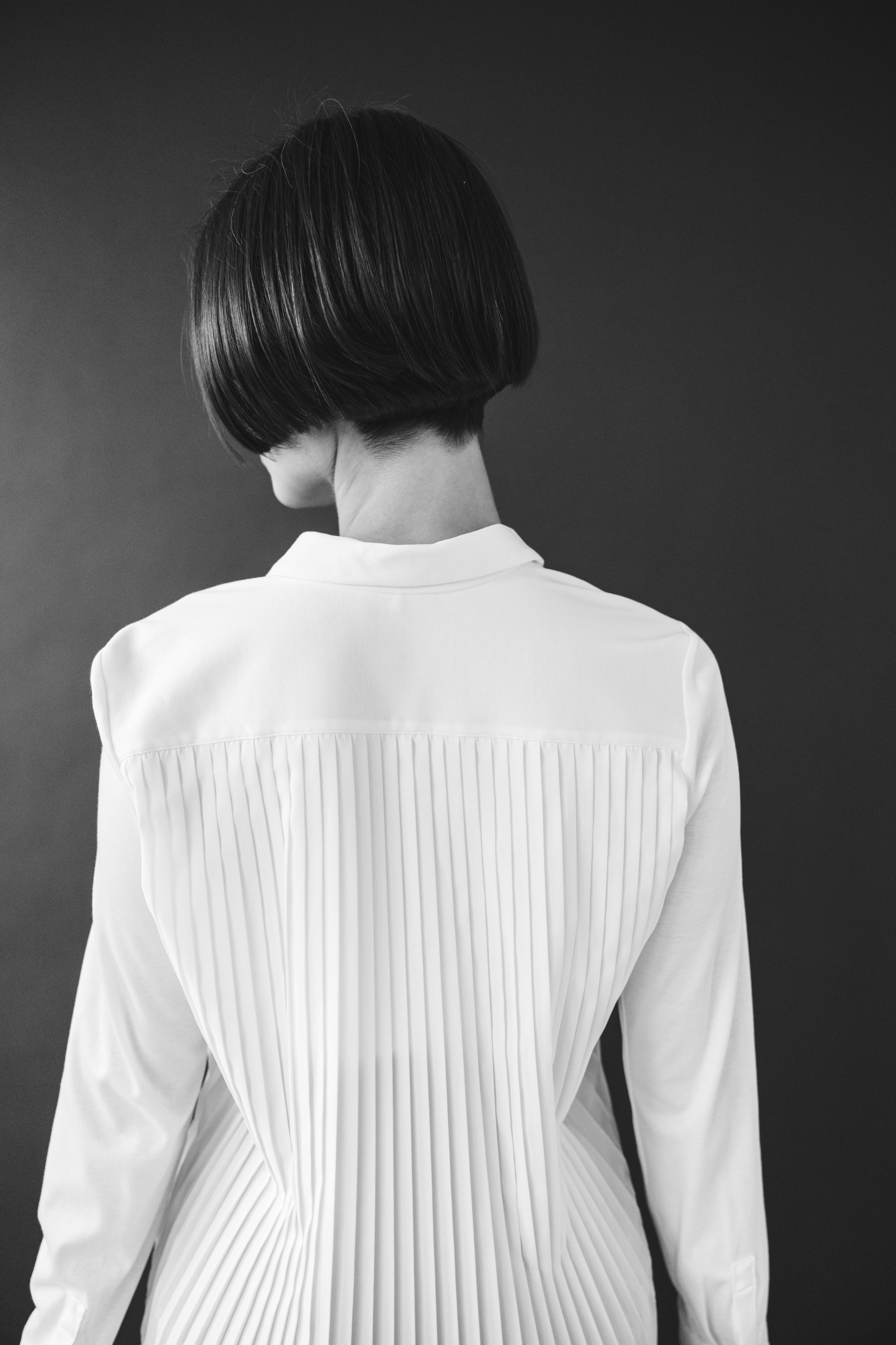 Person with short bob haircut wearing a white pleated-back shirt facing away against a dark background.