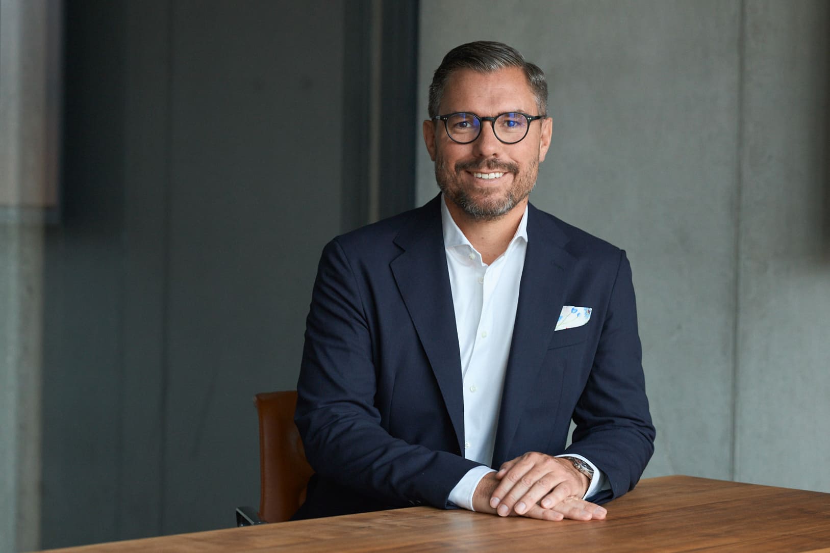 Man with beard and glasses wearing a navy blazer and white shirt sitting at a wooden table in an office.
