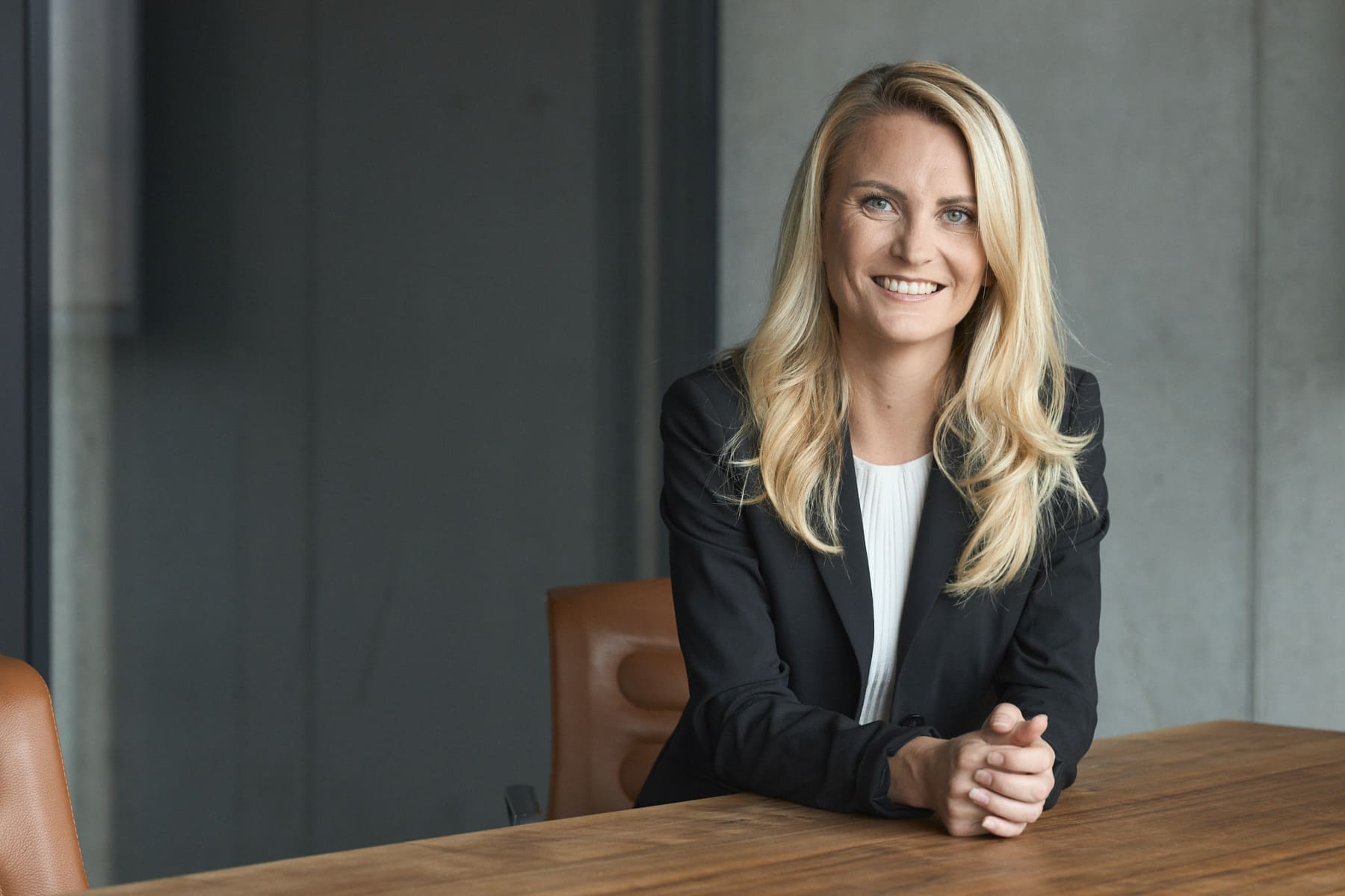 Smiling woman with long blonde hair in a black blazer sitting at a wooden conference table.