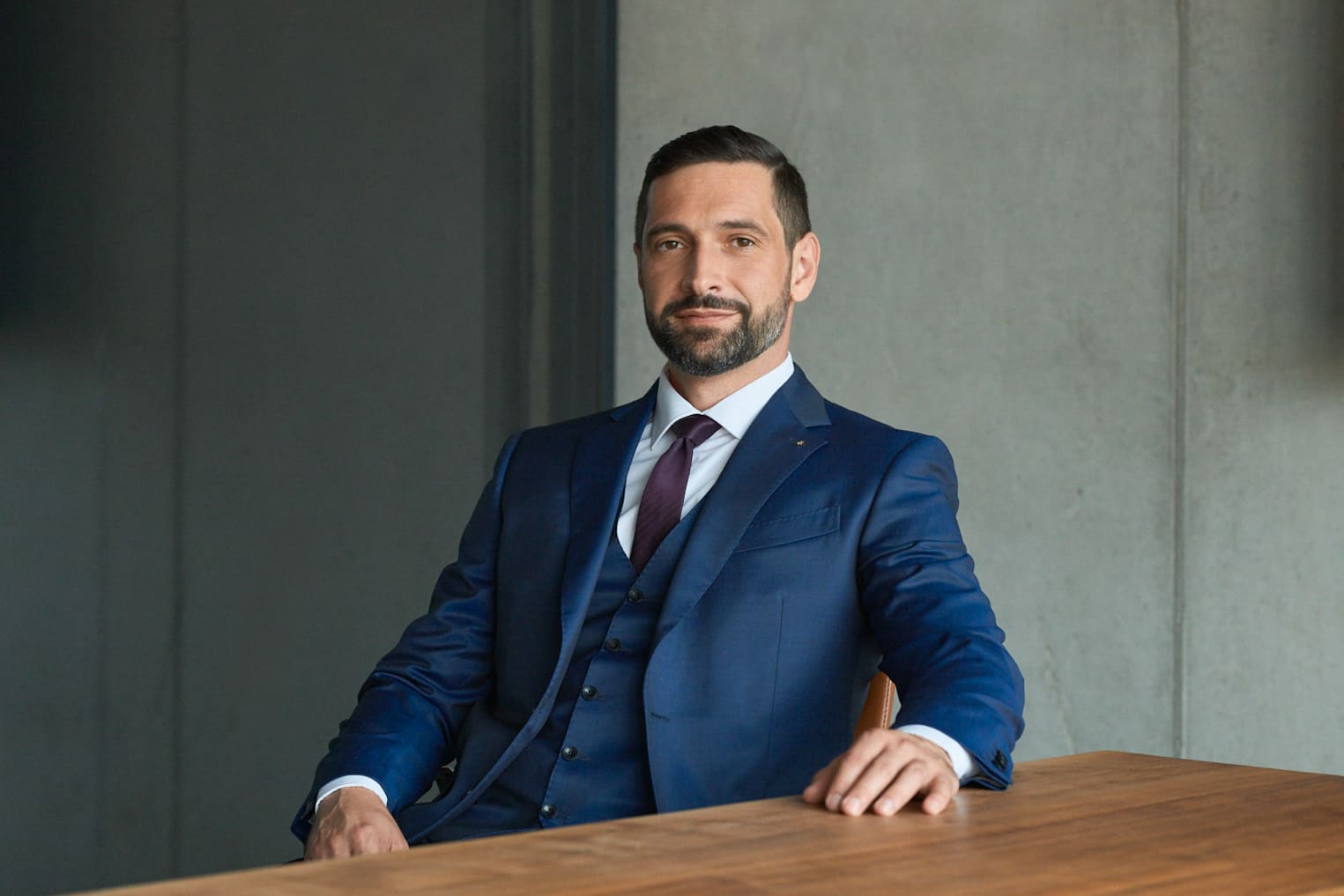 Man with beard wearing a blue suit and purple tie sitting at a wooden table with a neutral expression.