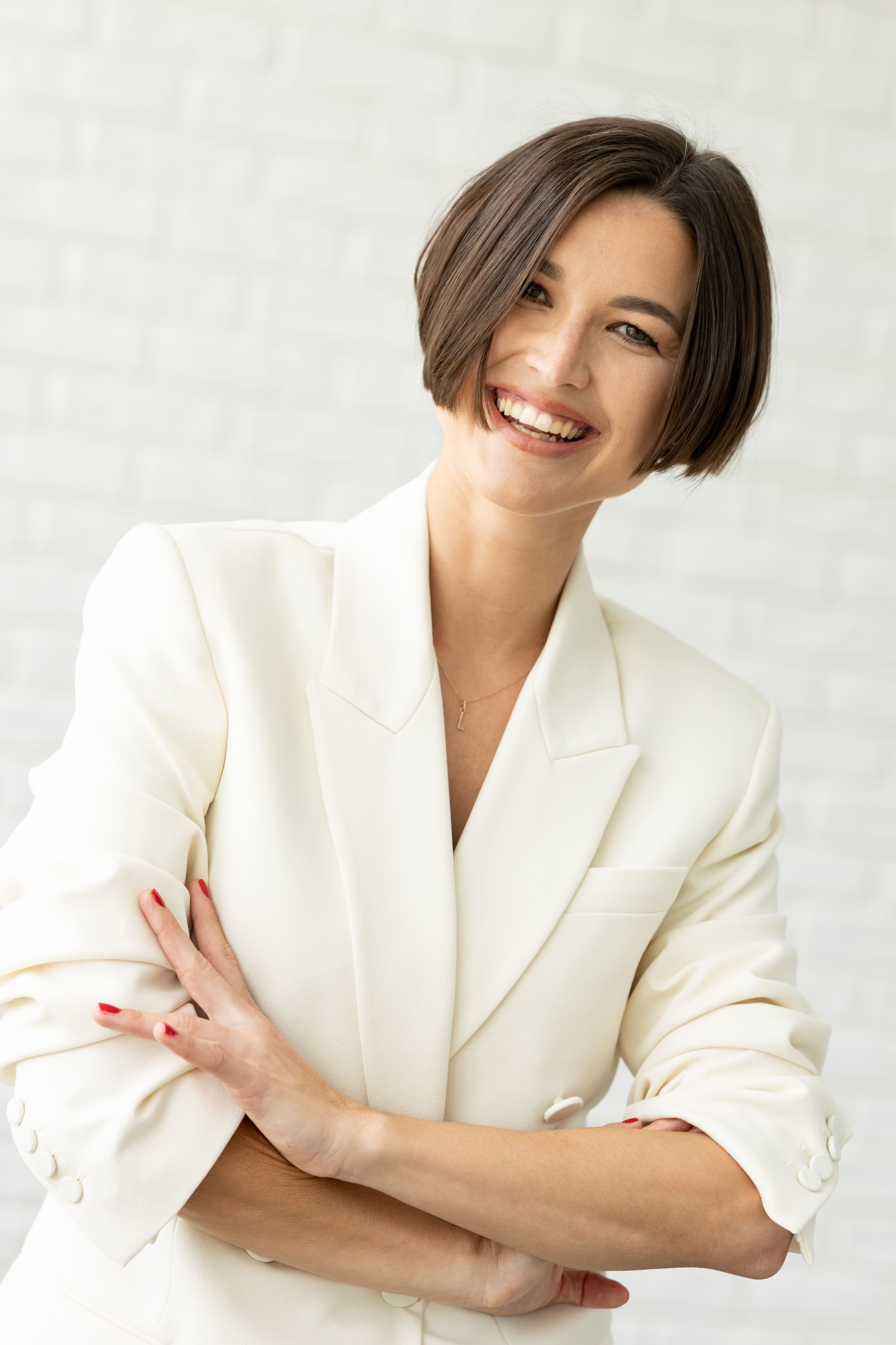 Smiling woman with short brown hair wearing a white blazer standing with folded arms against a light brick wall.