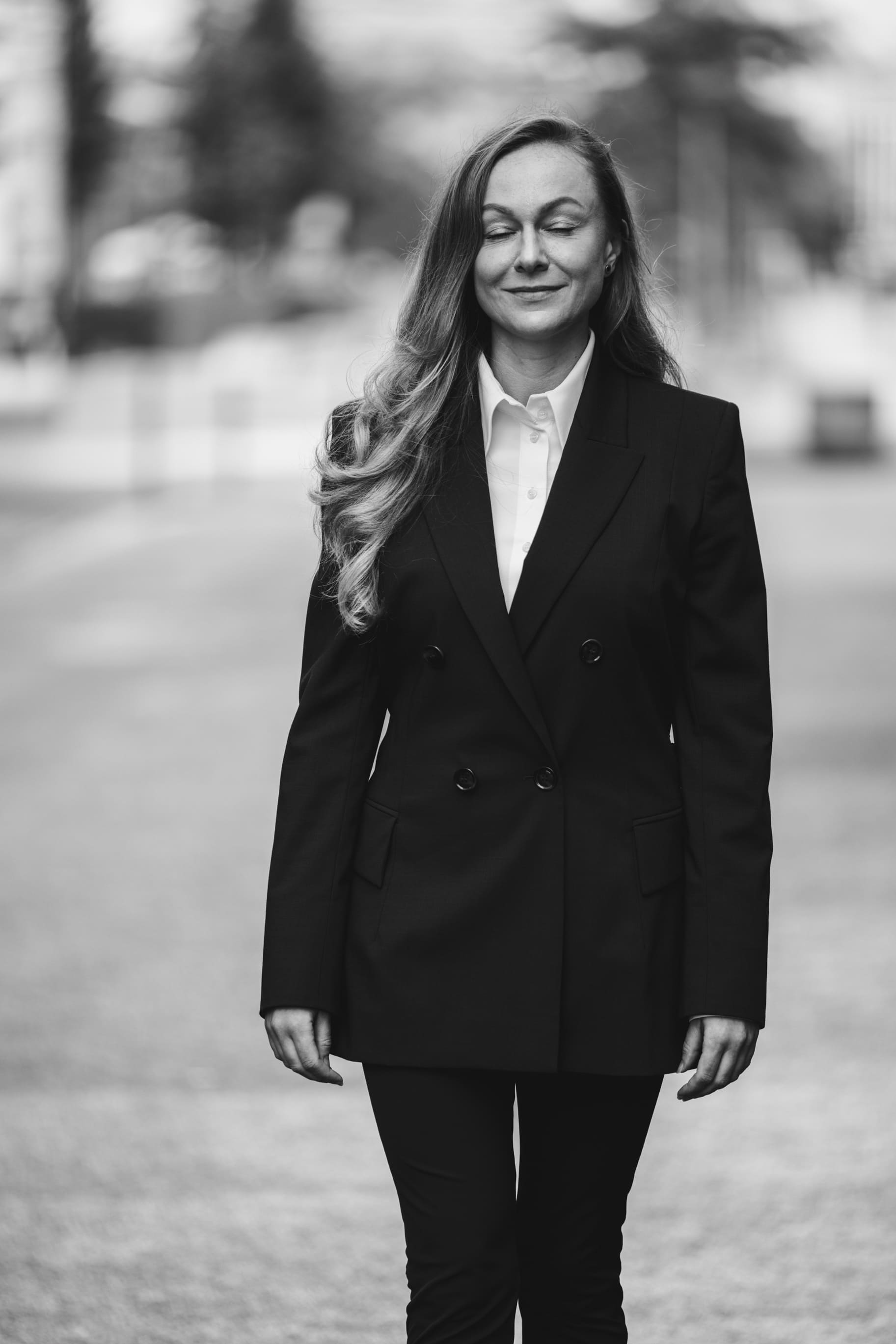 Woman in a black blazer with long hair walking outdoors with eyes closed and a slight smile.
