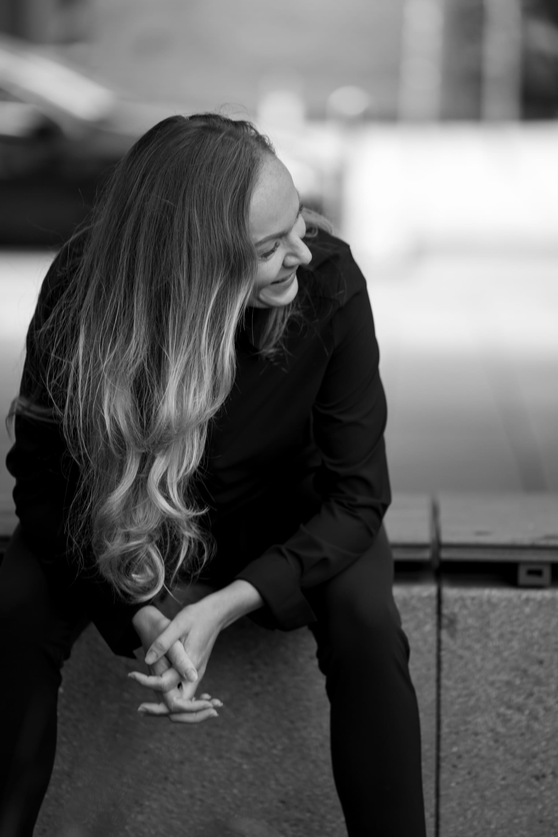 Black and white photo of a woman with long hair sitting on a stone ledge, smiling and looking to her left.