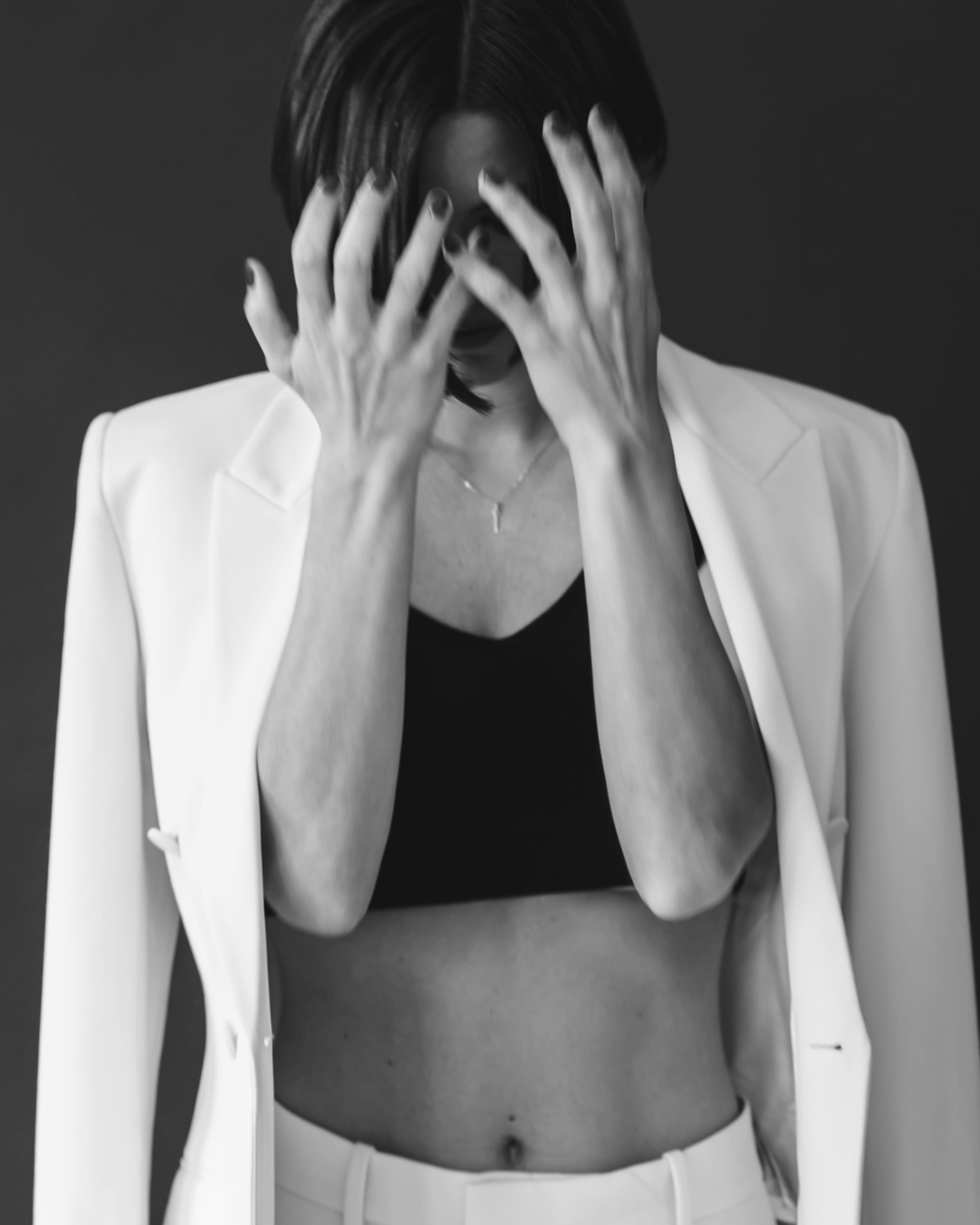 Black and white photo of a woman in a white blazer and pants with a black crop top, covering her face with her hands.