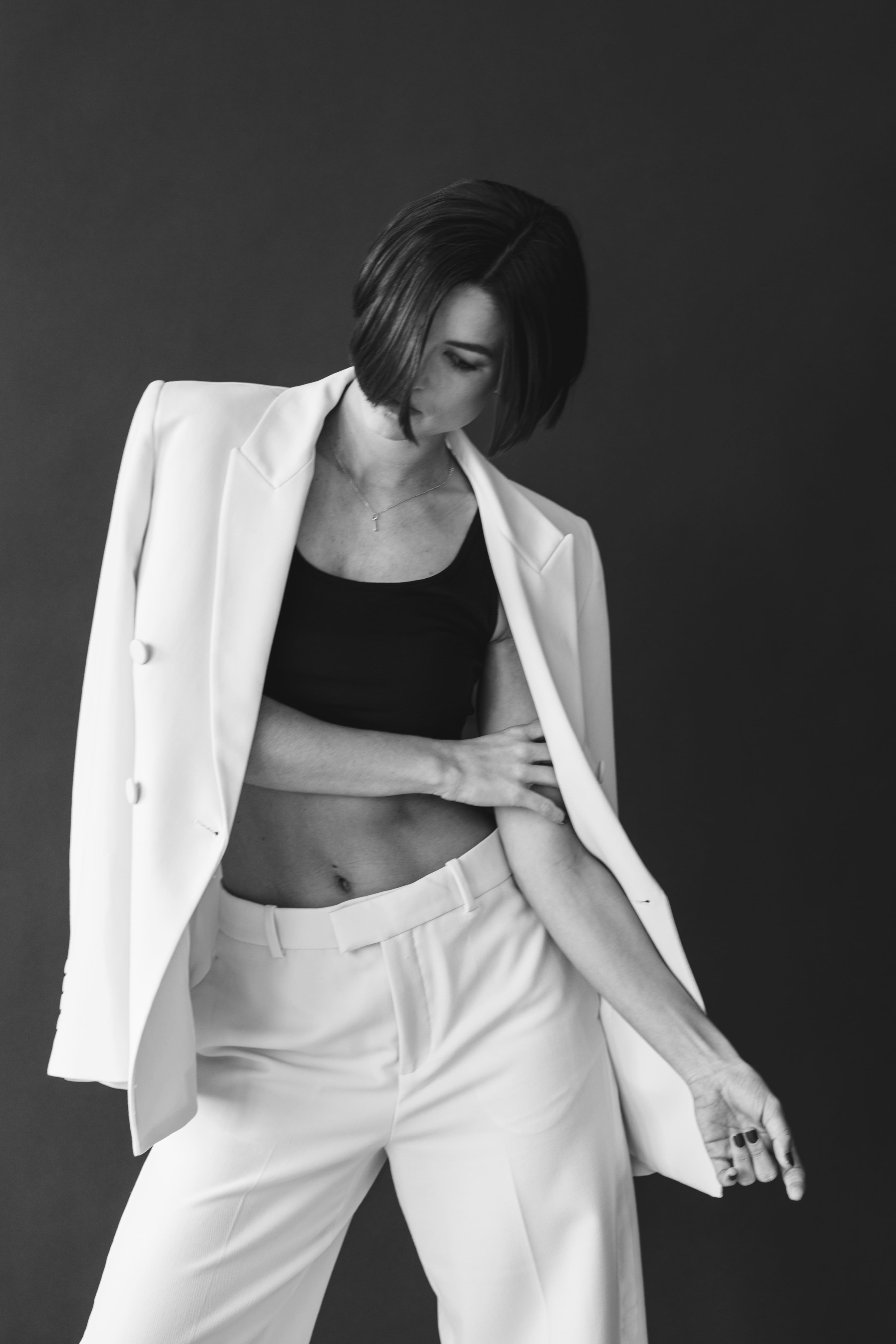 Black and white photo of a woman with short hair wearing a white blazer draped over her shoulders, a black crop top, and white pants against a dark background.