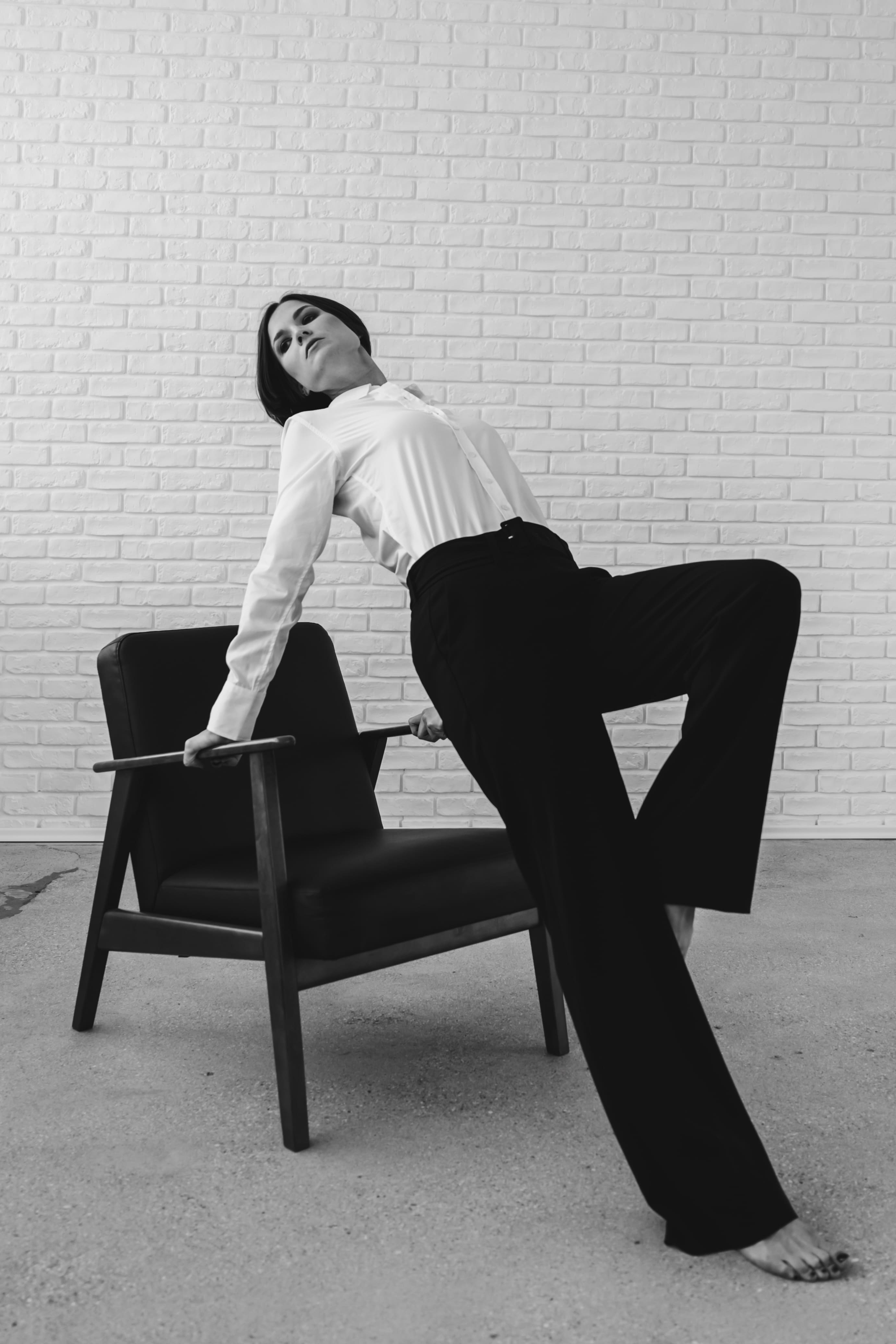 Black and white photo of a barefoot woman in black pants and a white shirt leaning back on a chair against a white brick wall.