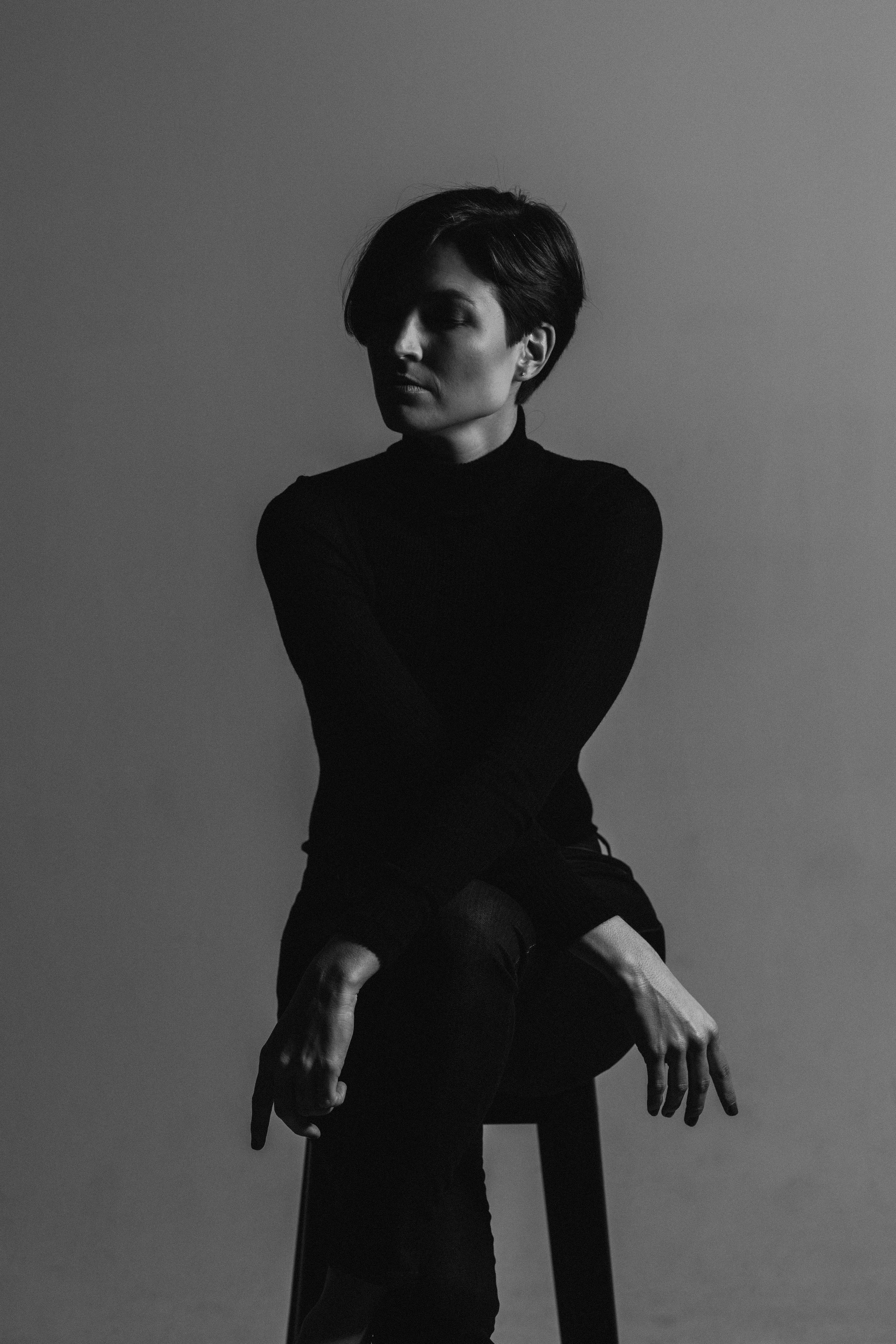 Black and white portrait of a person with short hair sitting on a stool, looking to the side with arms resting on their legs.