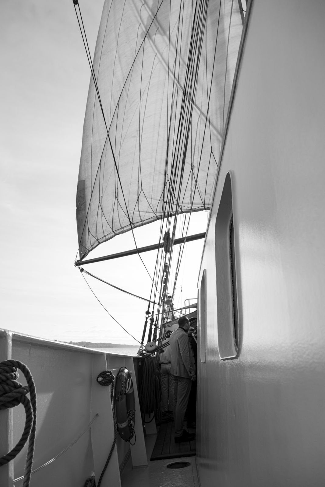 Black-and-white photo showing a man in a suit standing on the deck of a sailboat under a large sail with rigging lines.