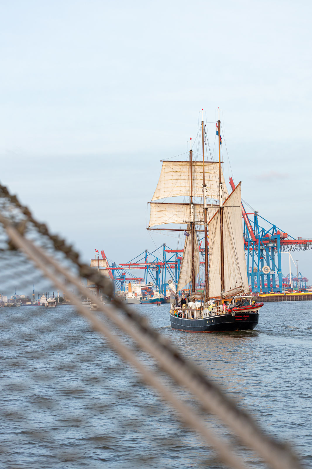 Sailboat with beige sails sailing on water with blue and red port cranes in the background.