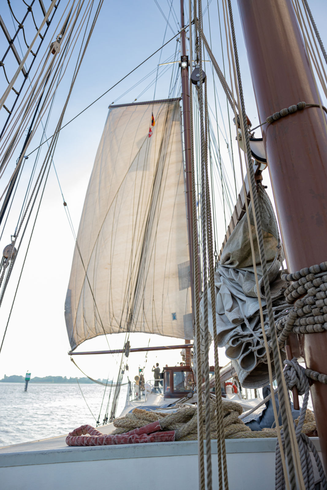 View along the deck of a sailboat with sails hoisted and ropes arranged, sailing on a calm body of water near the shore.