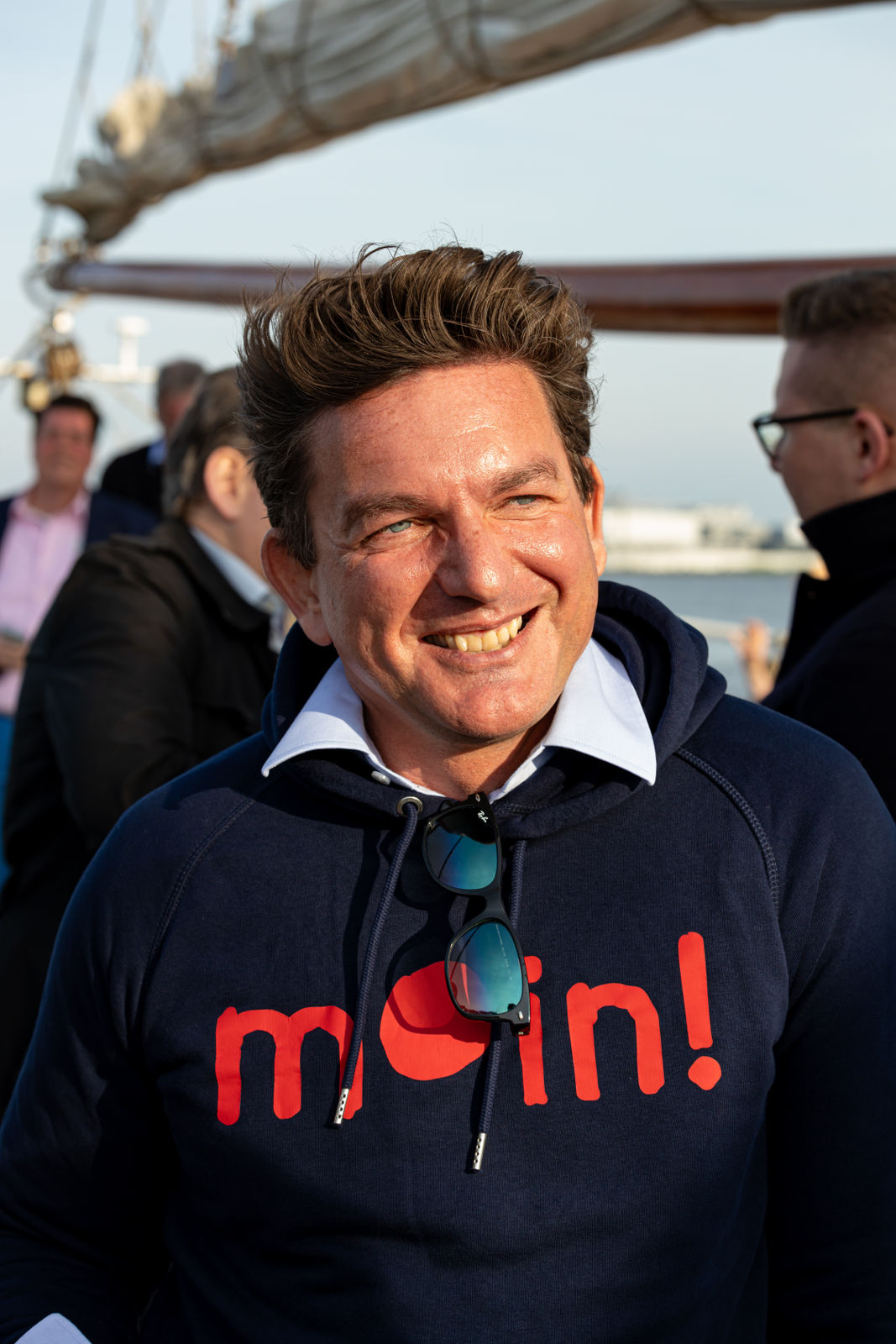 Smiling man with styled hair wearing a navy hoodie with red text and sunglasses hanging from the collar, standing outdoors near water.