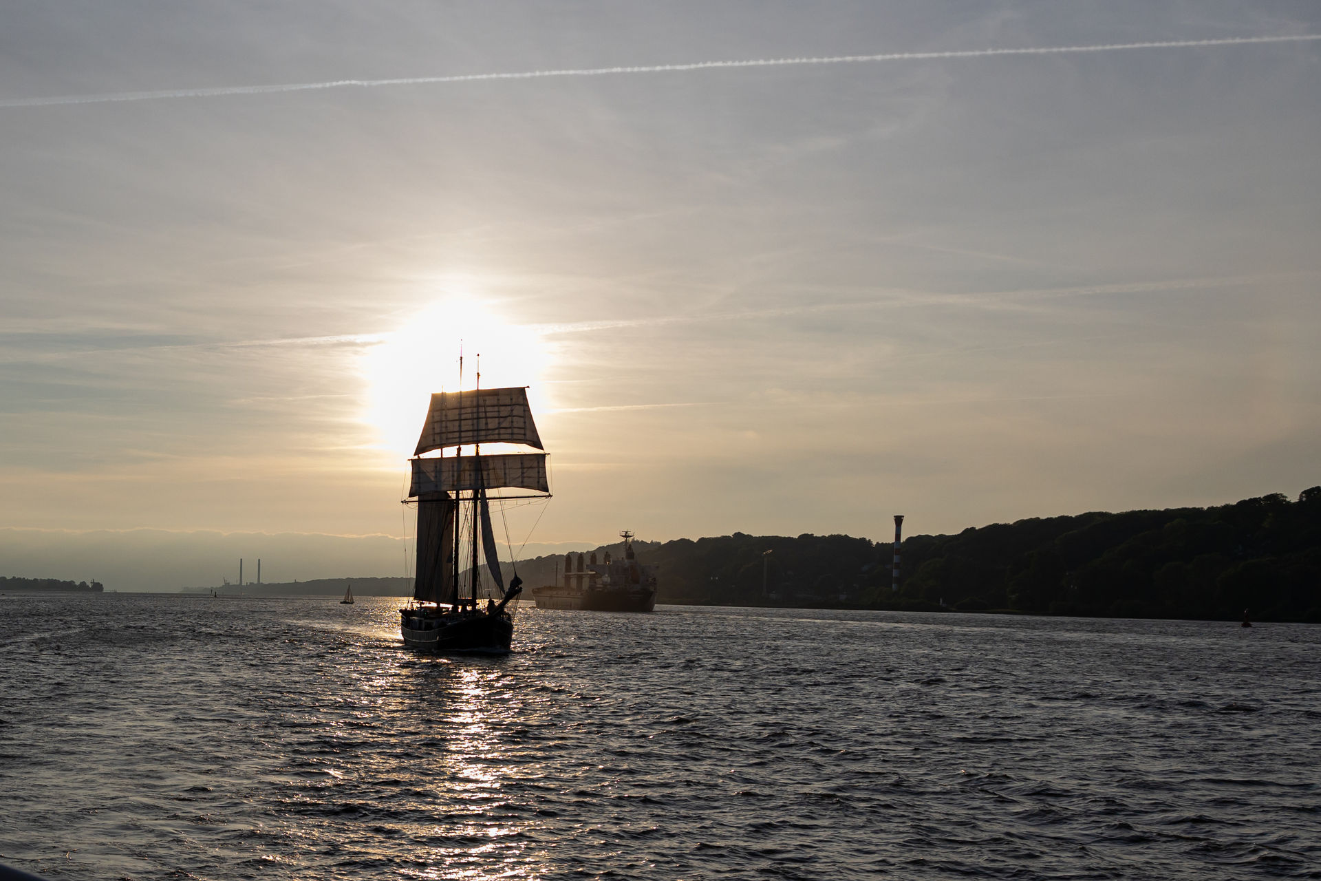 Sailboat with raised sails silhouetted against the setting sun on a calm body of water, with another ship and forested land in the background.