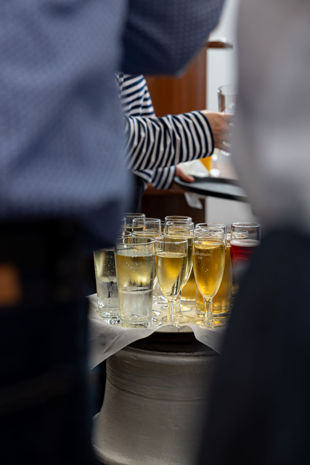 Tray of assorted drinks including glasses of beer and sparkling water being carried by a person in a striped shirt.