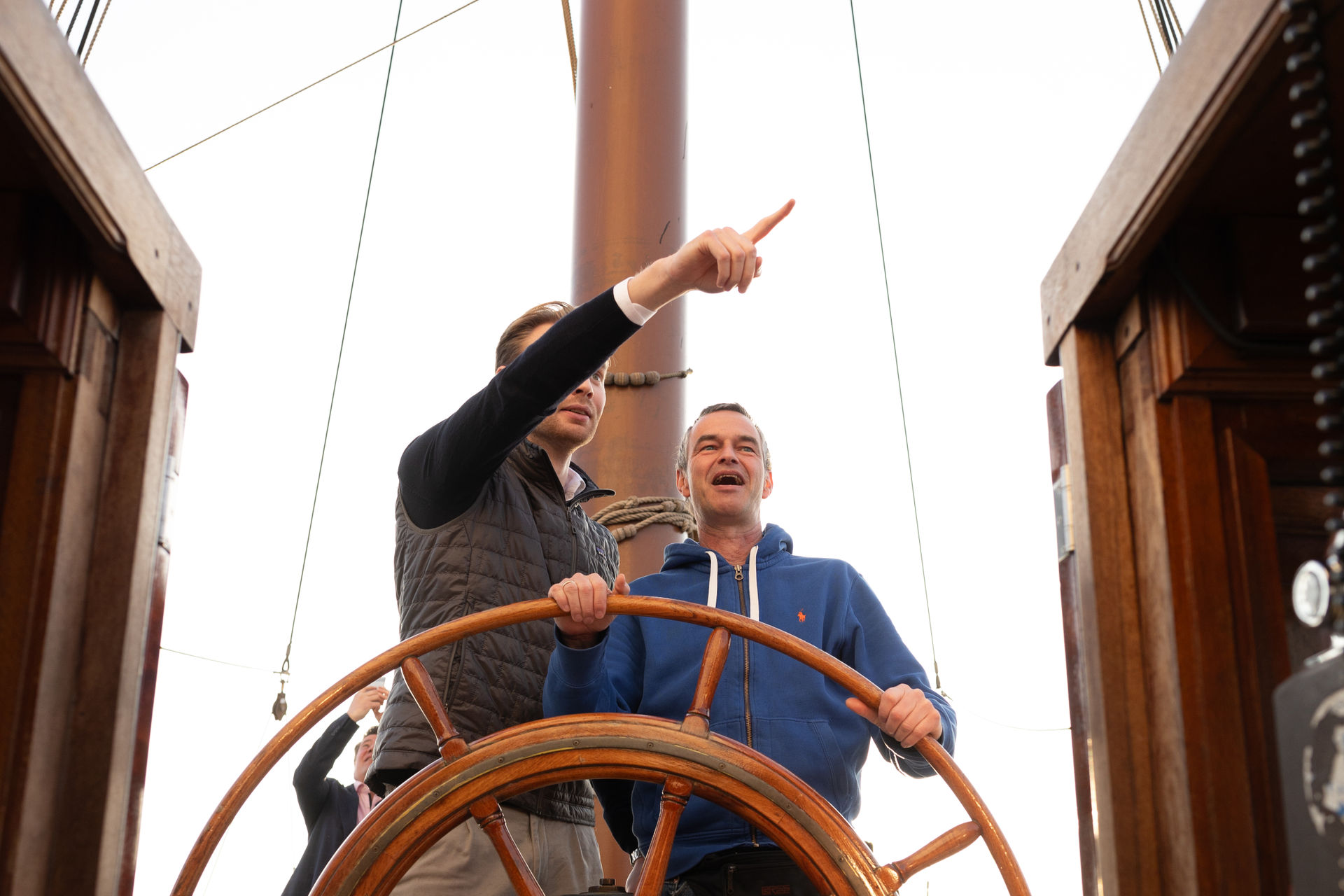 Two men steering a wooden ship wheel, one pointing ahead and the other smiling, with a tall mast in the background.