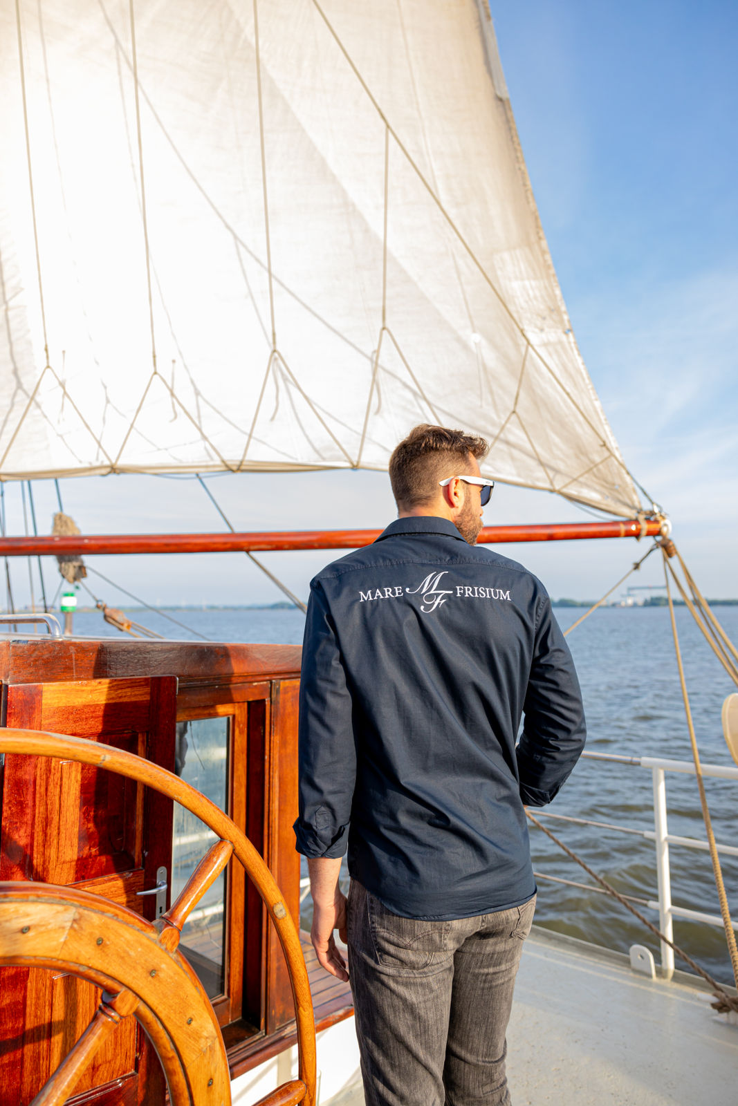 Man wearing sunglasses and a navy shirt with 'Mare Frisium' on the back standing on a sailboat under a large white sail on a sunny day.
