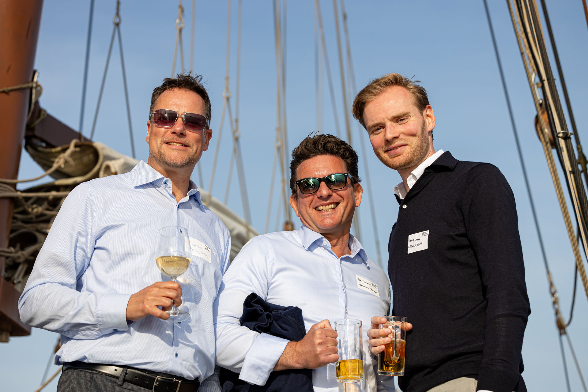 Three men smiling and holding drinks on a boat with rigging and a clear blue sky in the background.