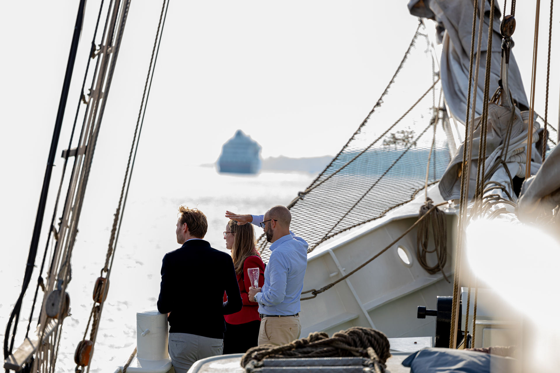 Three people standing on a sailboat deck looking out at the water with a large ship in the distance.