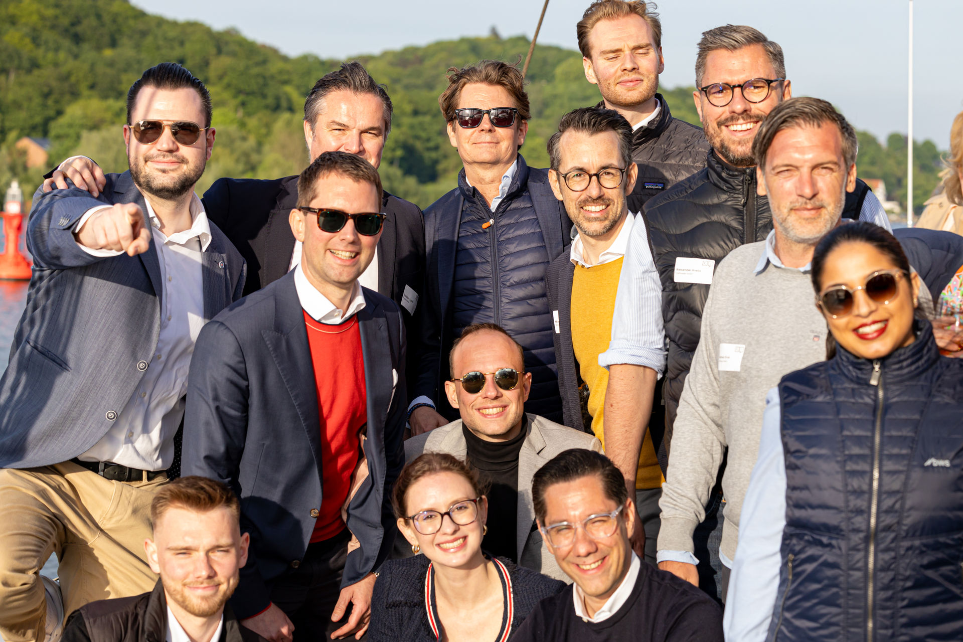 Group of smiling people outdoors in casual and business attire with trees and water in the background.