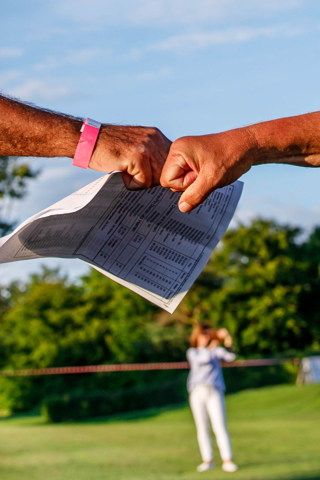Two people fist bumping outdoors, holding a paper with printed text, with a blurred person and greenery in the background.