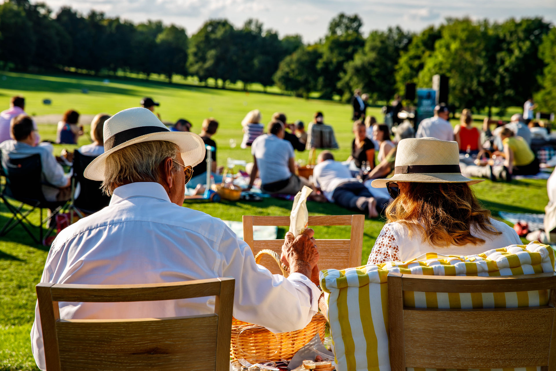 Older couple wearing hats sitting on wooden chairs with a picnic basket at an outdoor gathering on a sunny day.