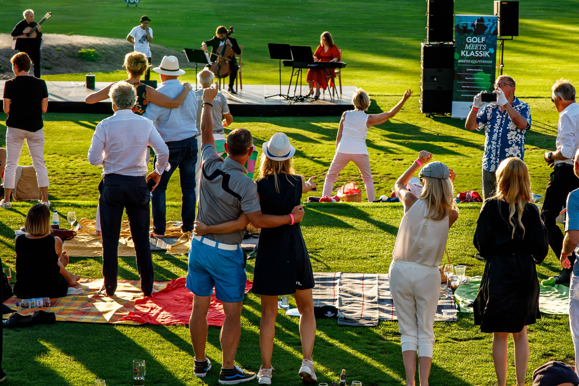 People standing and sitting on blankets on a lawn enjoying a live classical music performance outdoors during sunset.
