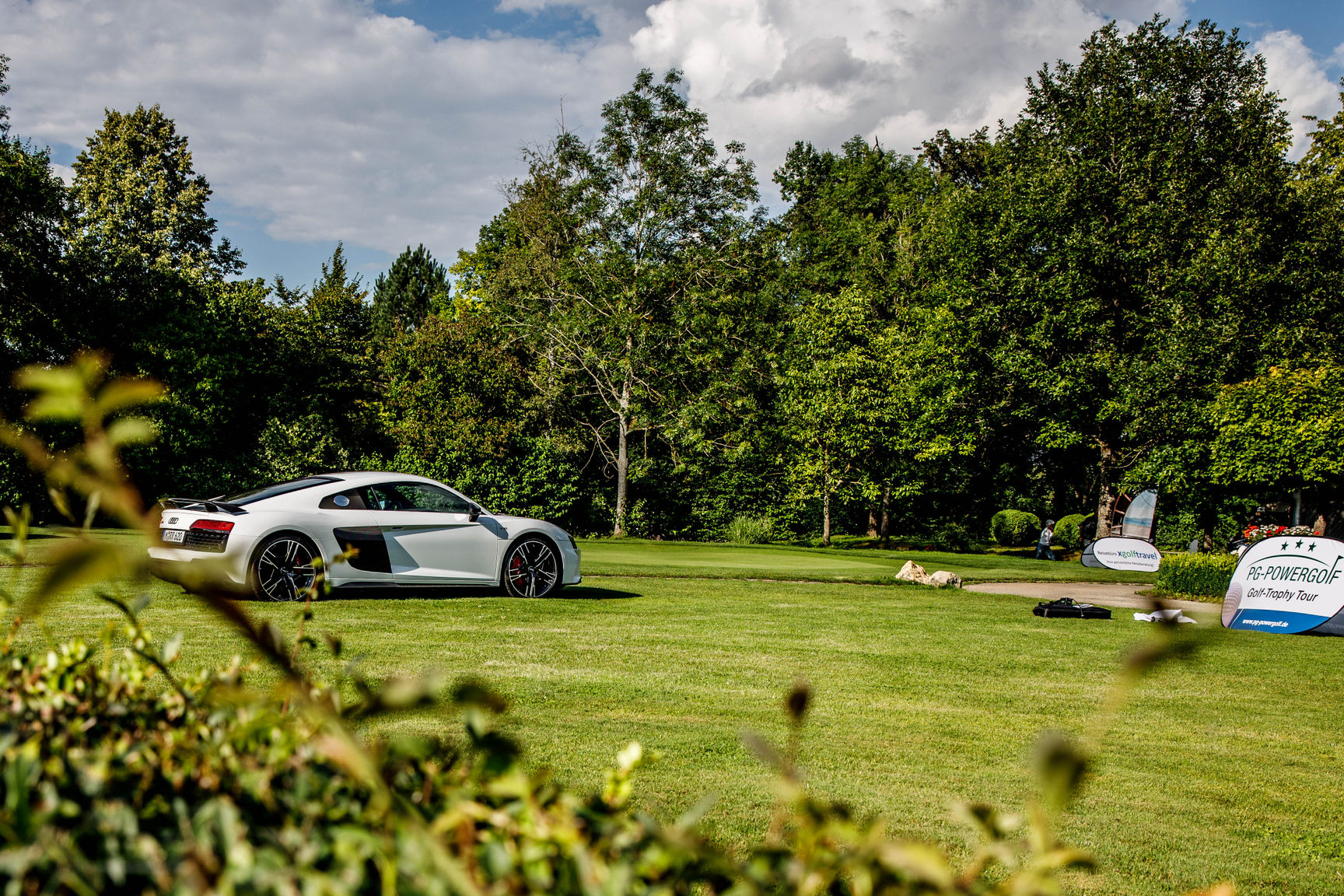 White sports car parked on a green golf course with trees and event banners in the background under a partly cloudy sky.