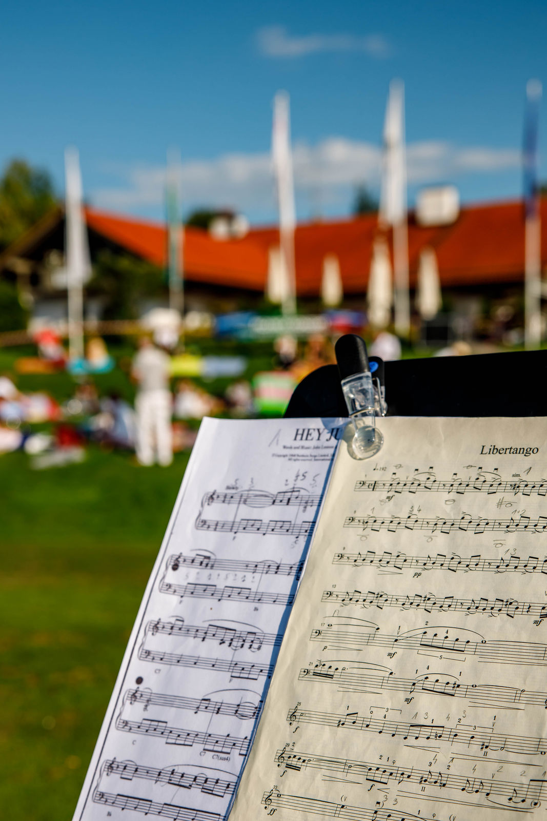 Sheet music for 'Libertango' and 'Hey Jude' on a music stand outdoors with people and a building with red roof in the blurred background.