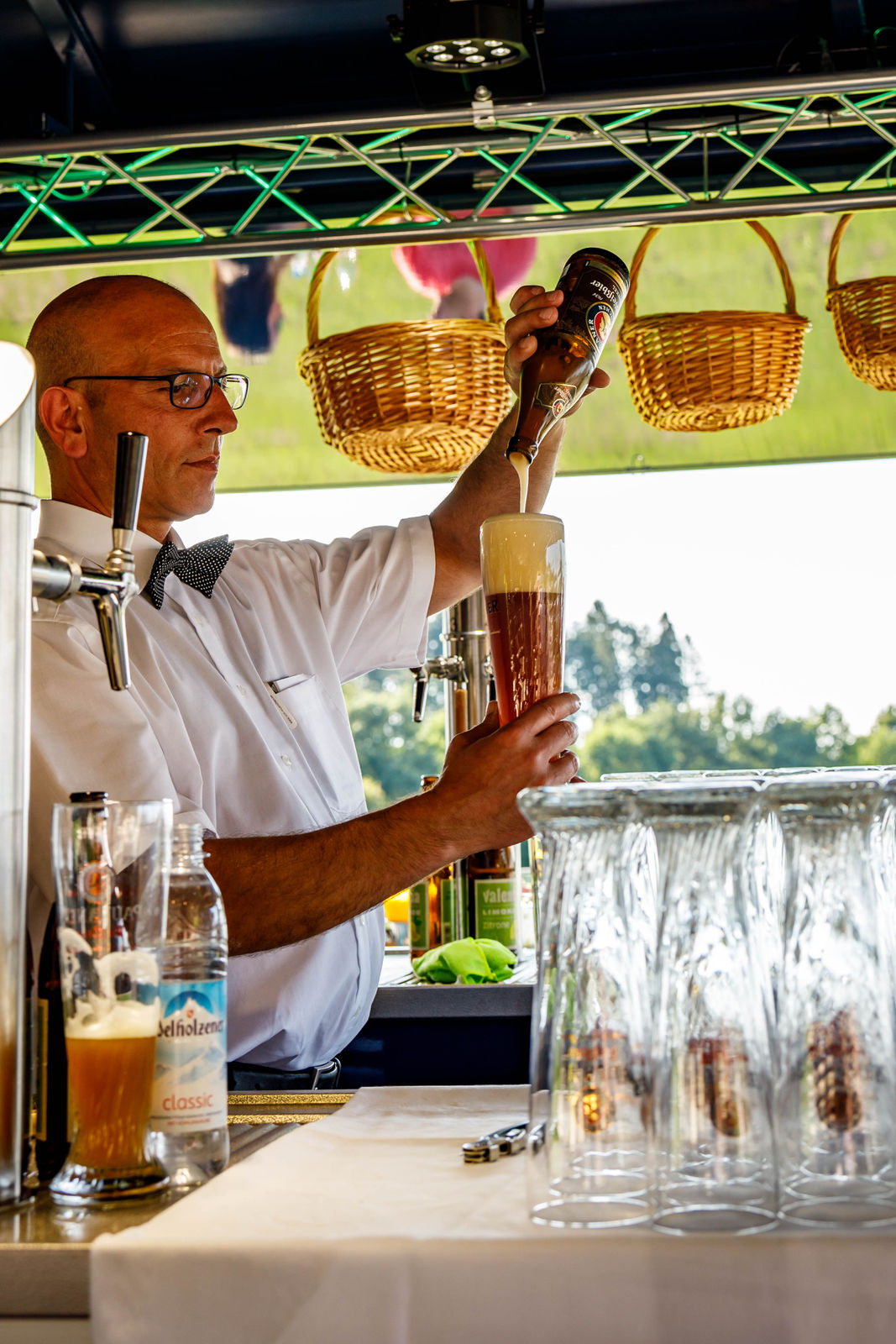 Bartender in white shirt and bow tie pouring beer from a bottle into a tall glass with foam head at an outdoor bar.