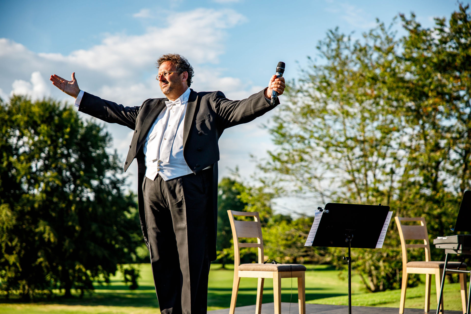 Man in a tuxedo holding a microphone with arms outstretched on an outdoor stage during daytime.