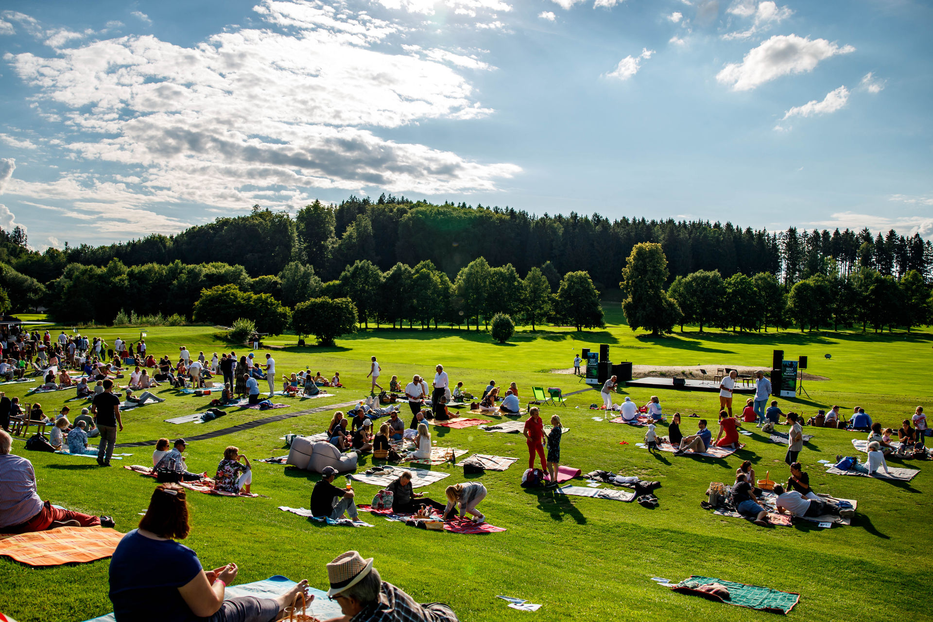 People sitting on blankets on a grassy field enjoying an outdoor event near a stage under a partly cloudy sky.