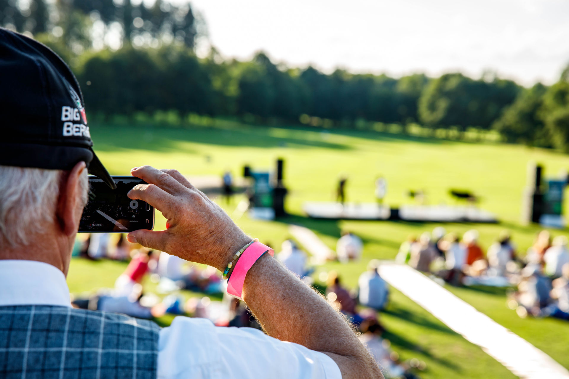 Man wearing a black cap and plaid vest recording an outdoor event on a smartphone.