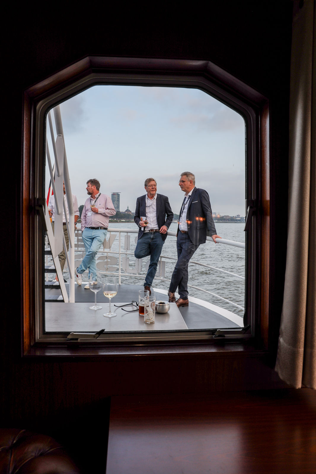 Three men in business casual attire conversing on a boat deck by the water, viewed through a wooden-framed window.