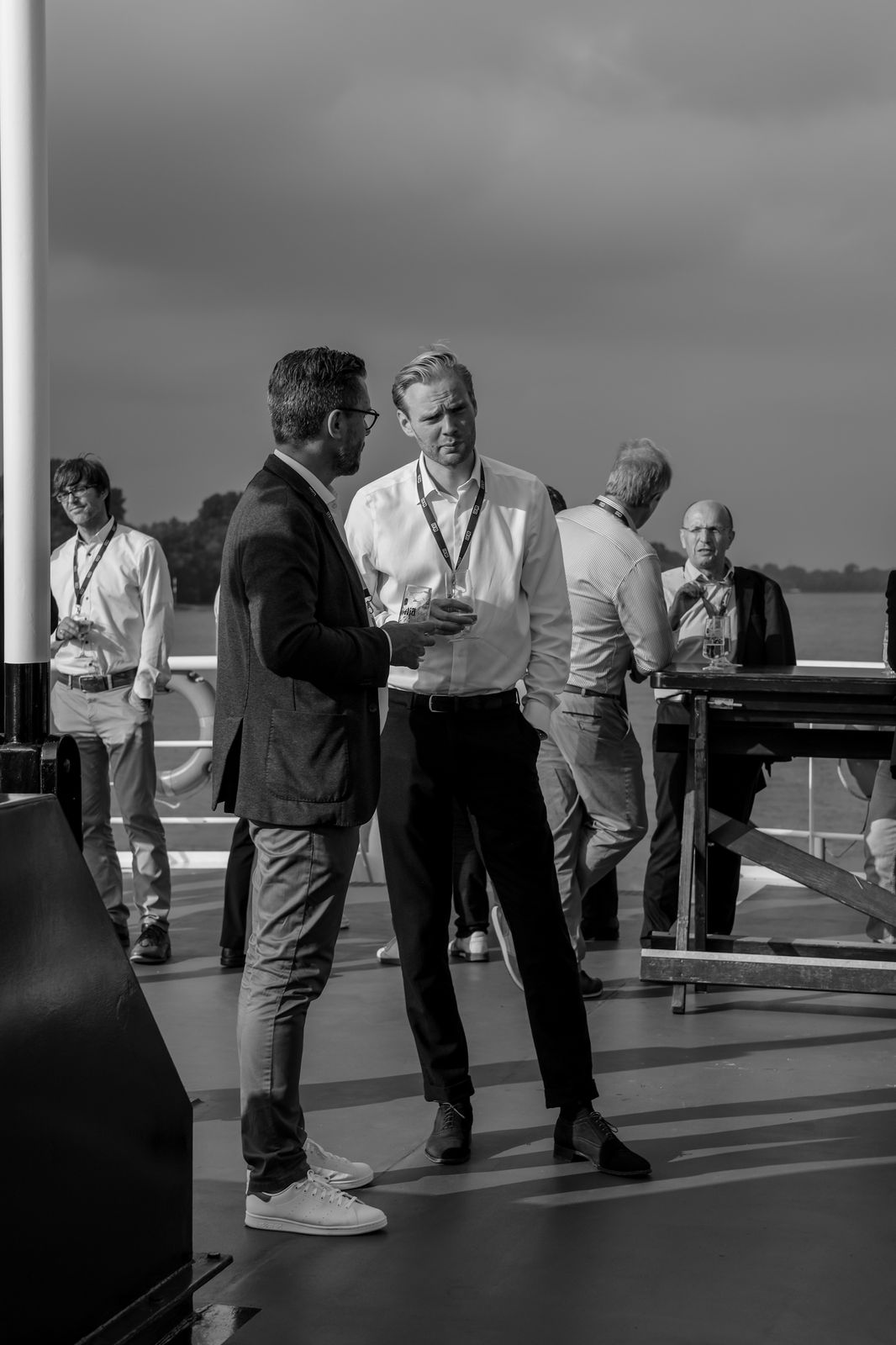 Group of men in business casual attire having conversations on an outdoor deck with a body of water in the background.
