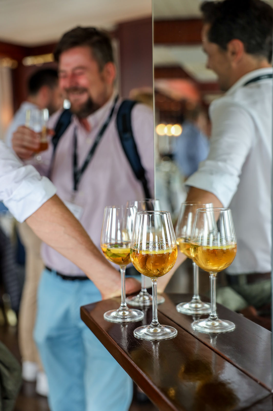 Three glasses of a golden drink on a wooden shelf with blurred men socializing in the background.