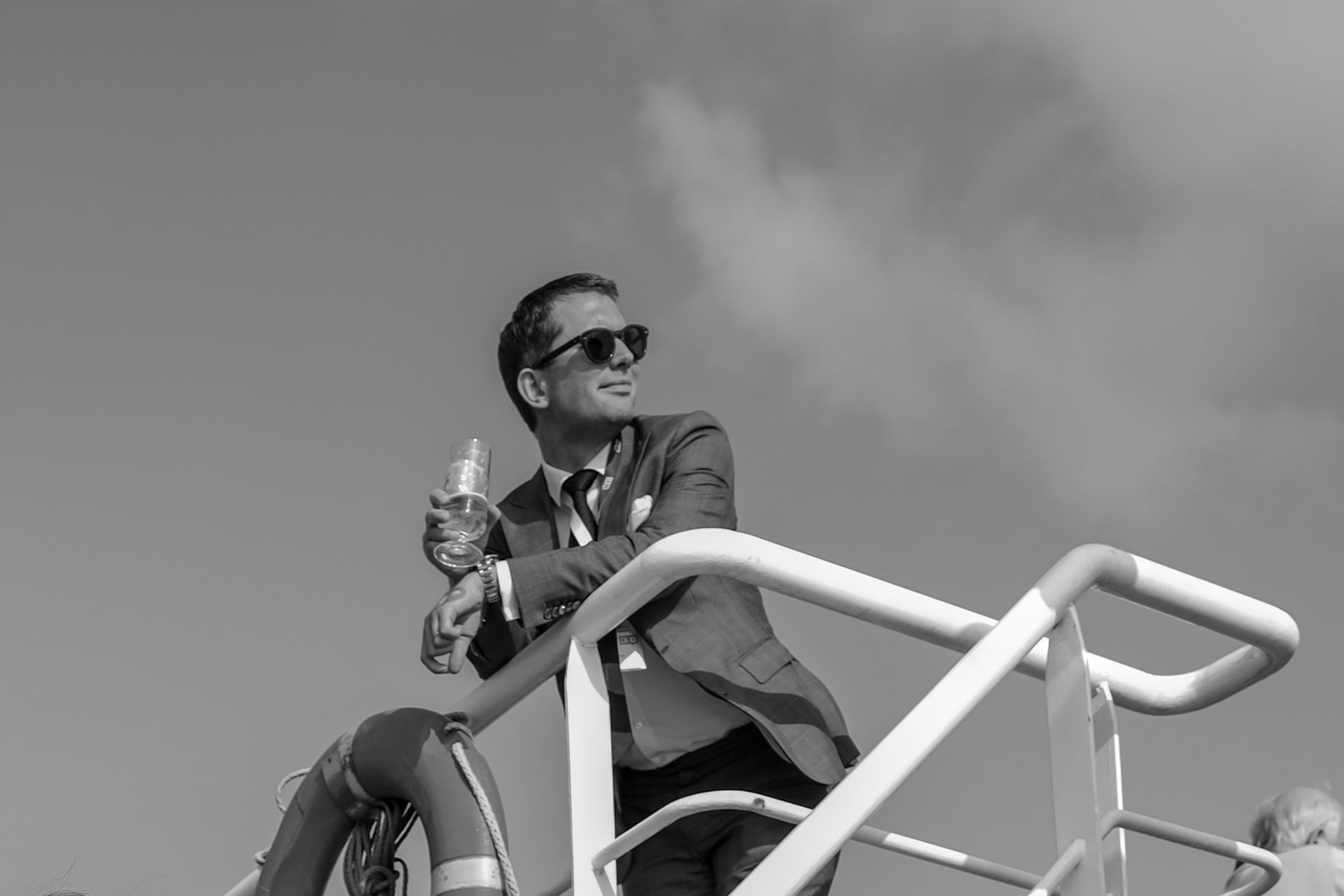 Man in a suit and sunglasses leaning on a railing holding a glass, looking into the distance outdoors.