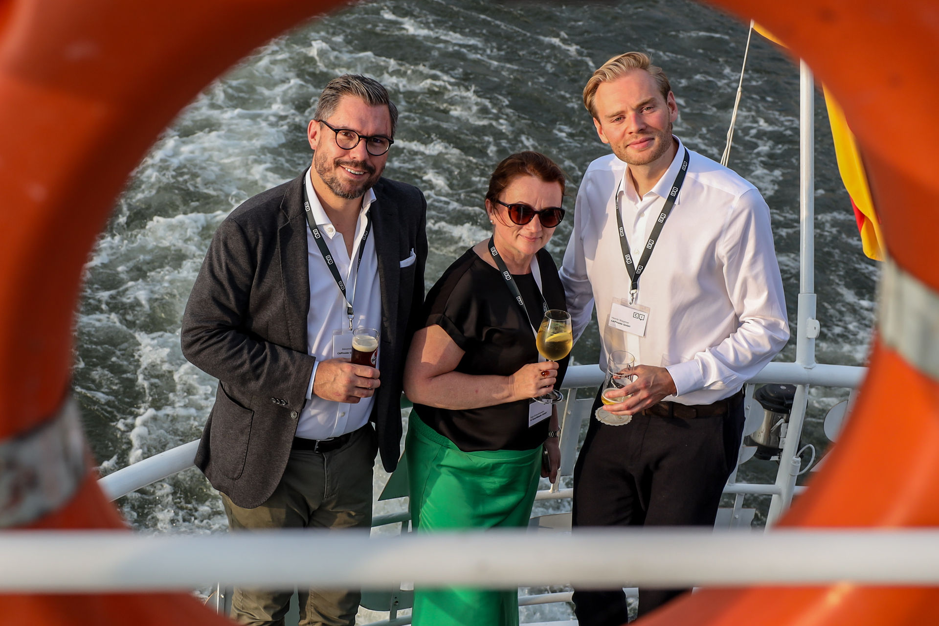 Three people with name badges standing on a boat deck holding drinks, framed by an orange lifebuoy with water in the background.