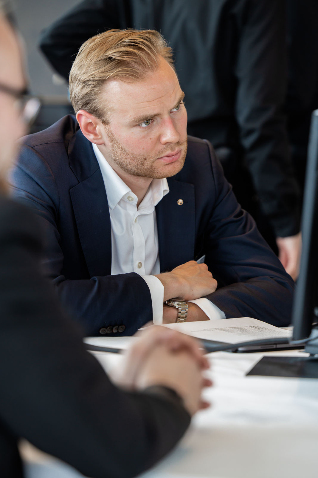 Man in a navy suit and white shirt attentively looking at a computer screen during a meeting.